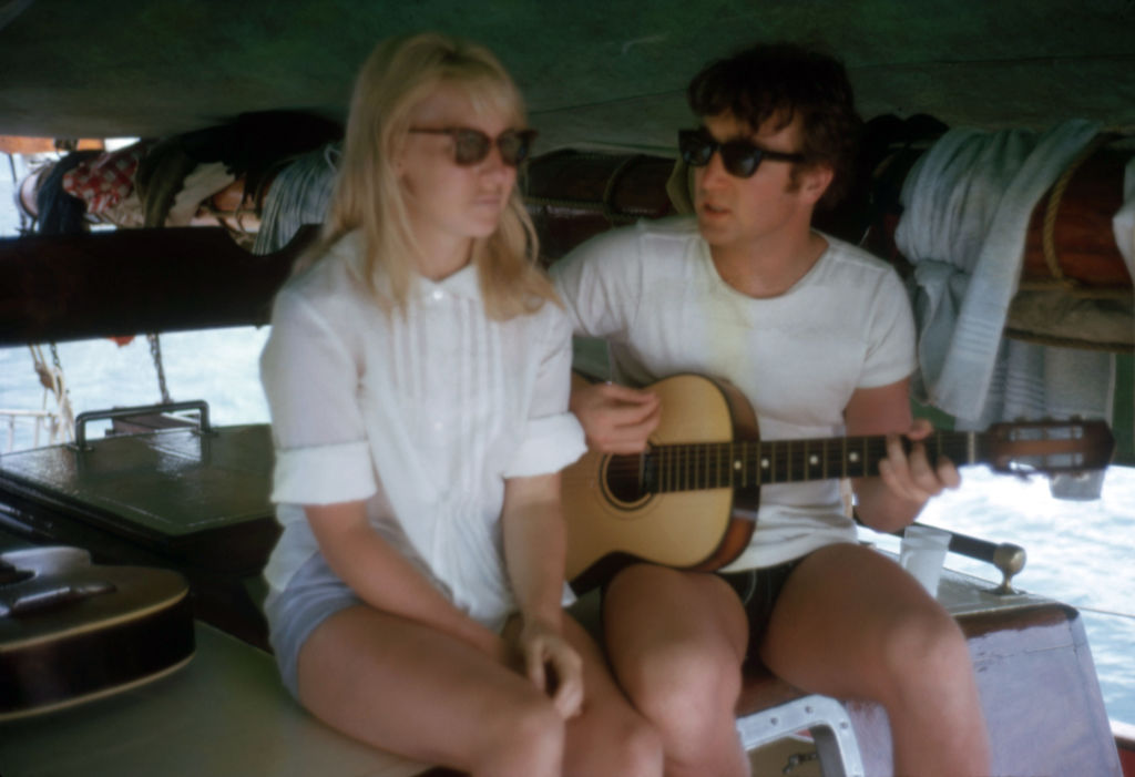 Cynthia Lennon sitting next to John Lennon as he plays guitar.