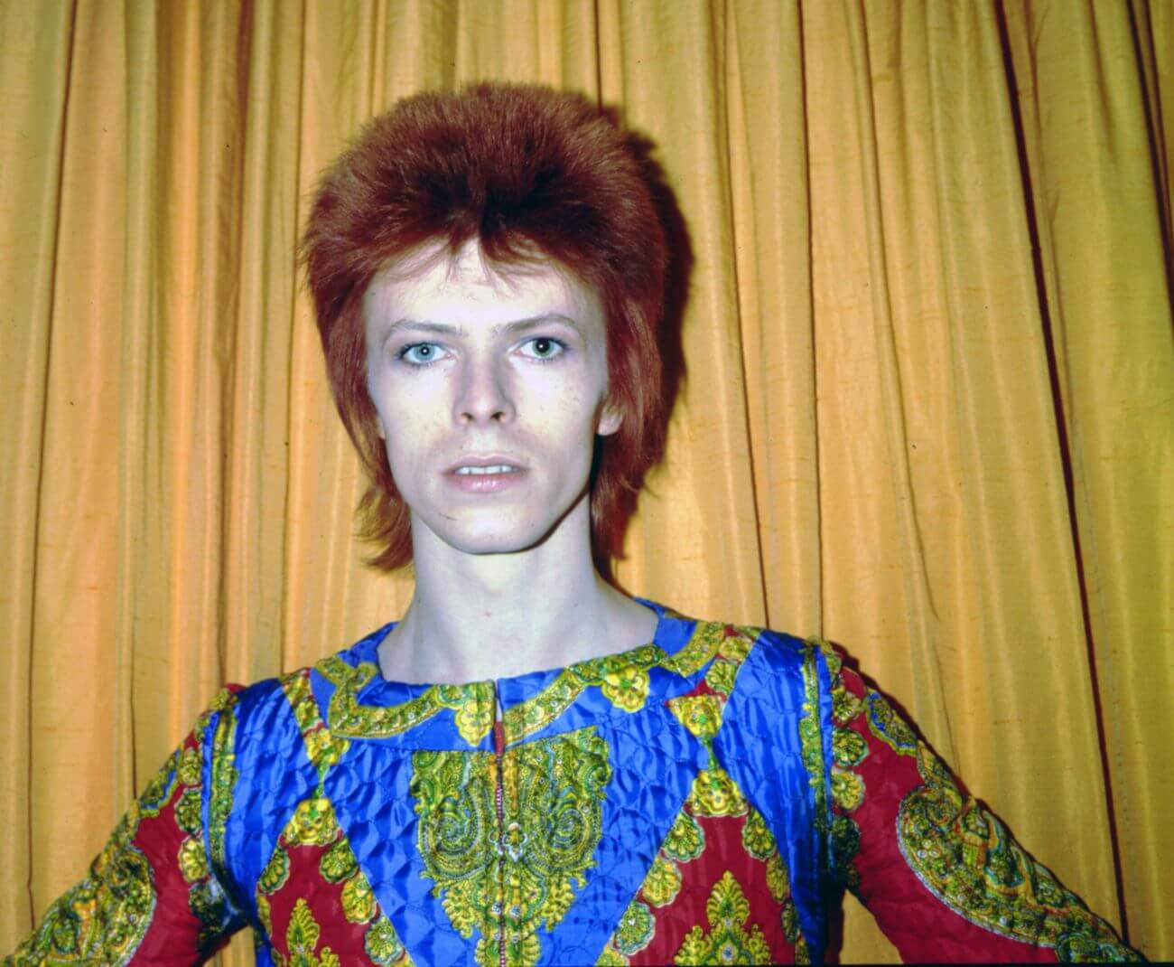 David Bowie wears a patterned shirt and stands with his hands on his hips.