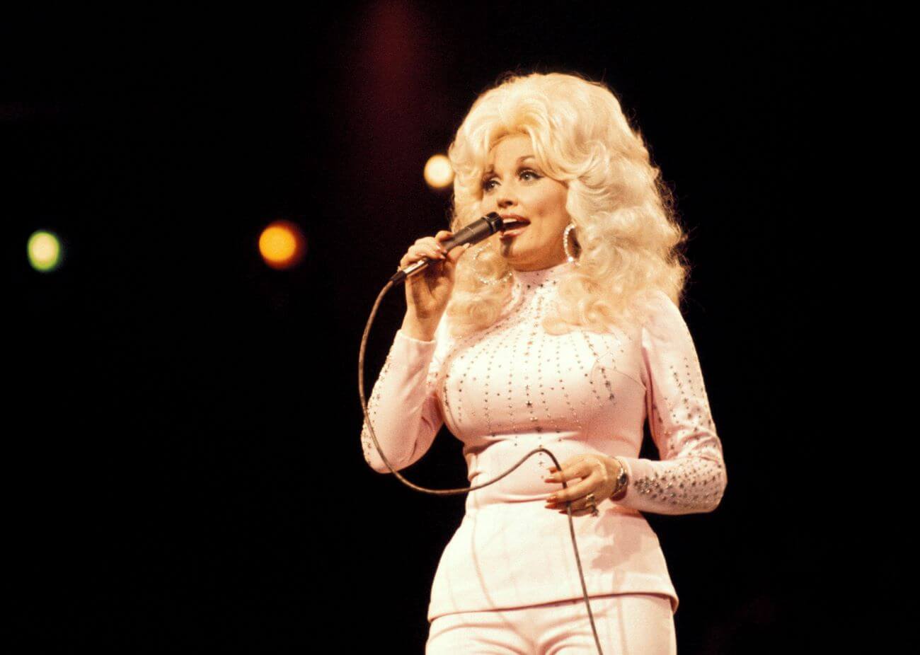 Dolly Parton wears a white outfit and holds a microphone.