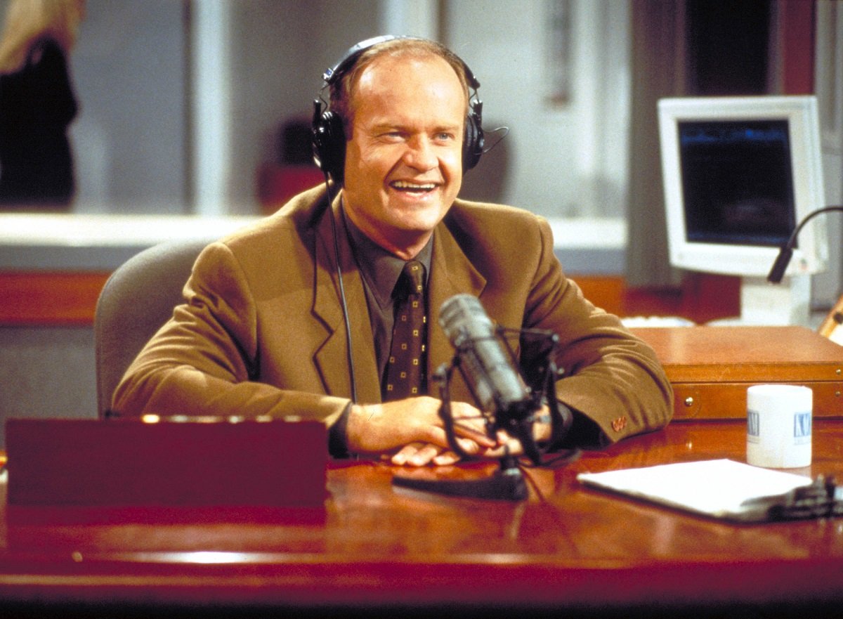Kelsey Grammer as Frasier Crane, wears a brown jacket while sitting at his radio studio desk in an episode of 'Frasier'