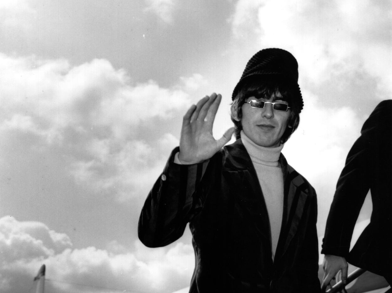George Harrison wears a dark coat, hat, and sunglasses as he waves while boarding a plane in 1966.