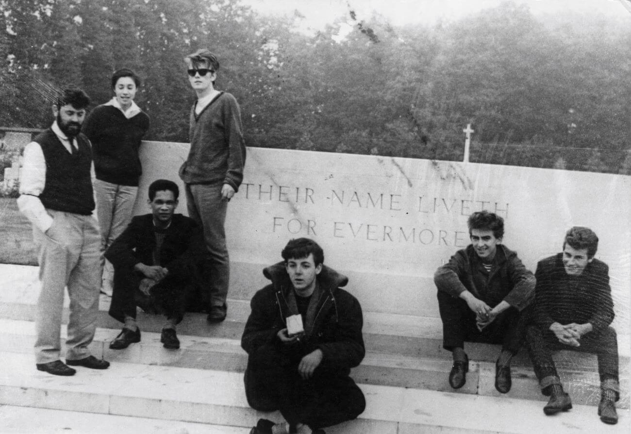 A black and white picture of Allan Williams, Beryl Williams, Lord Woodbine, Stuart Sutcliffe, Paul McCartney, George Harrison sitting on steps together.