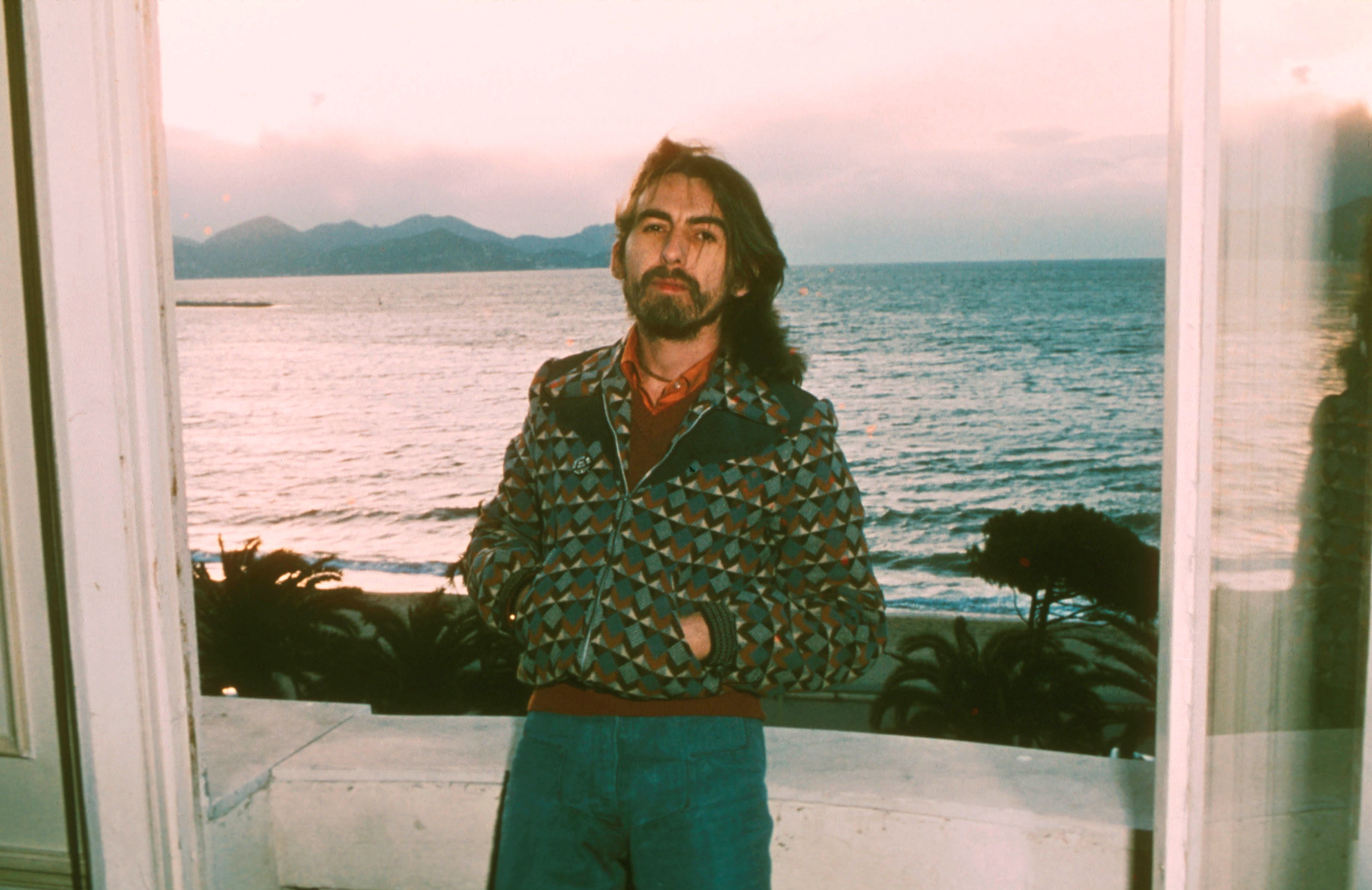 George Harrison poses with his hands in his jacket pockets in front of the ocean.