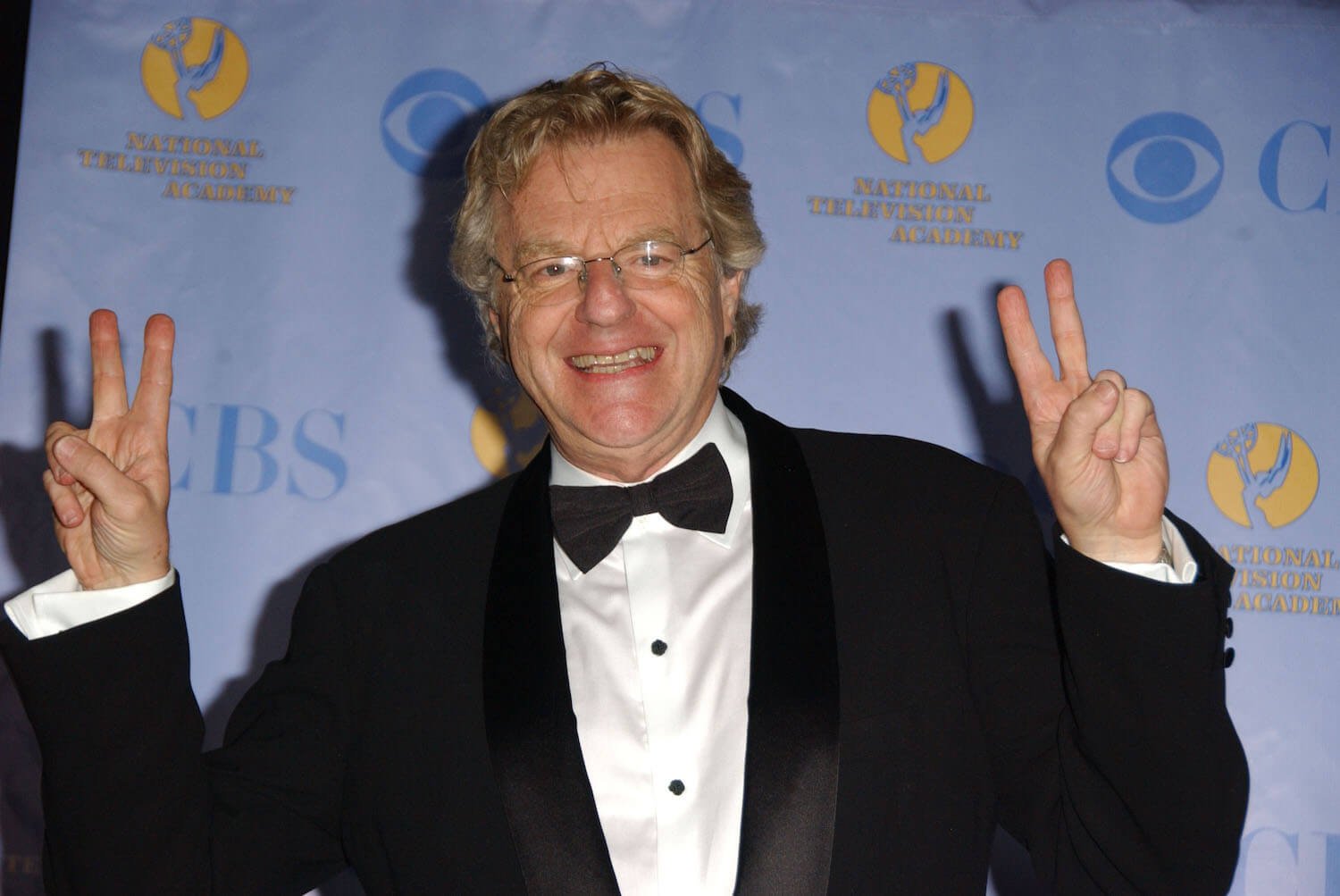 Jerry Springer holding his hands up in peace signs while wearing a suit. Jerry Springer died in 2023 with a high net worth.