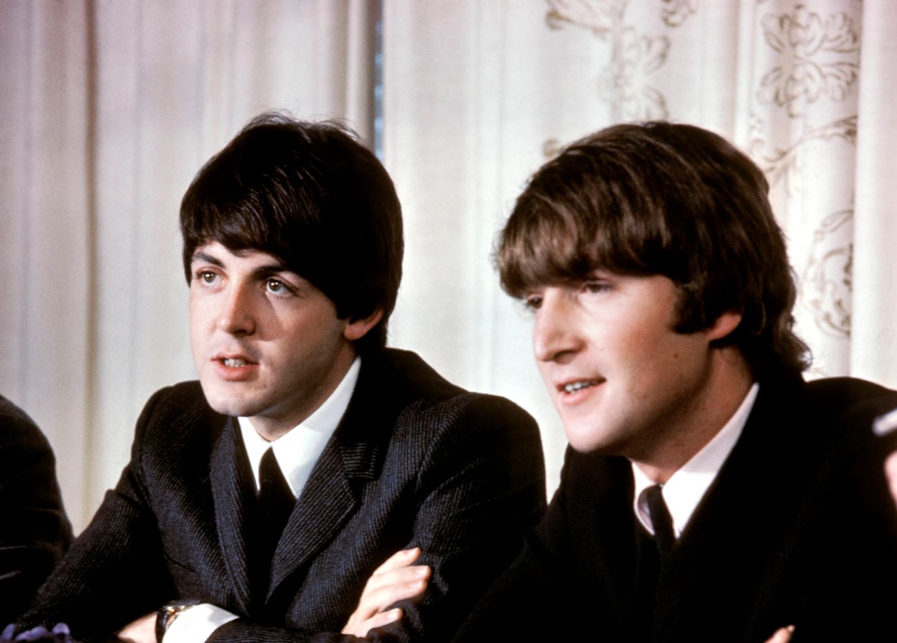 Paul McCartney and John Lennon wear suits and sit next to each other.
