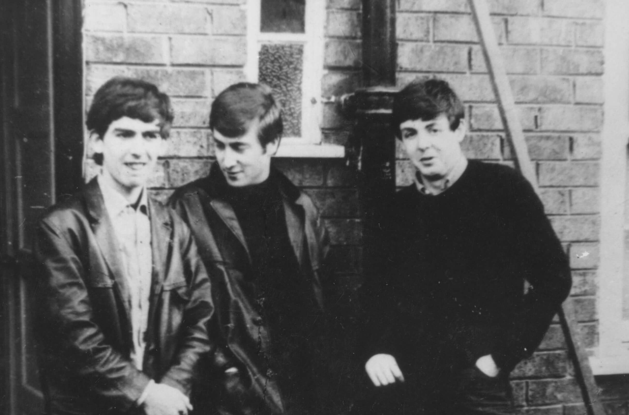 A black and white picture of George Harrison, John Lennon, and Paul McCartney leaning against a brick wall.