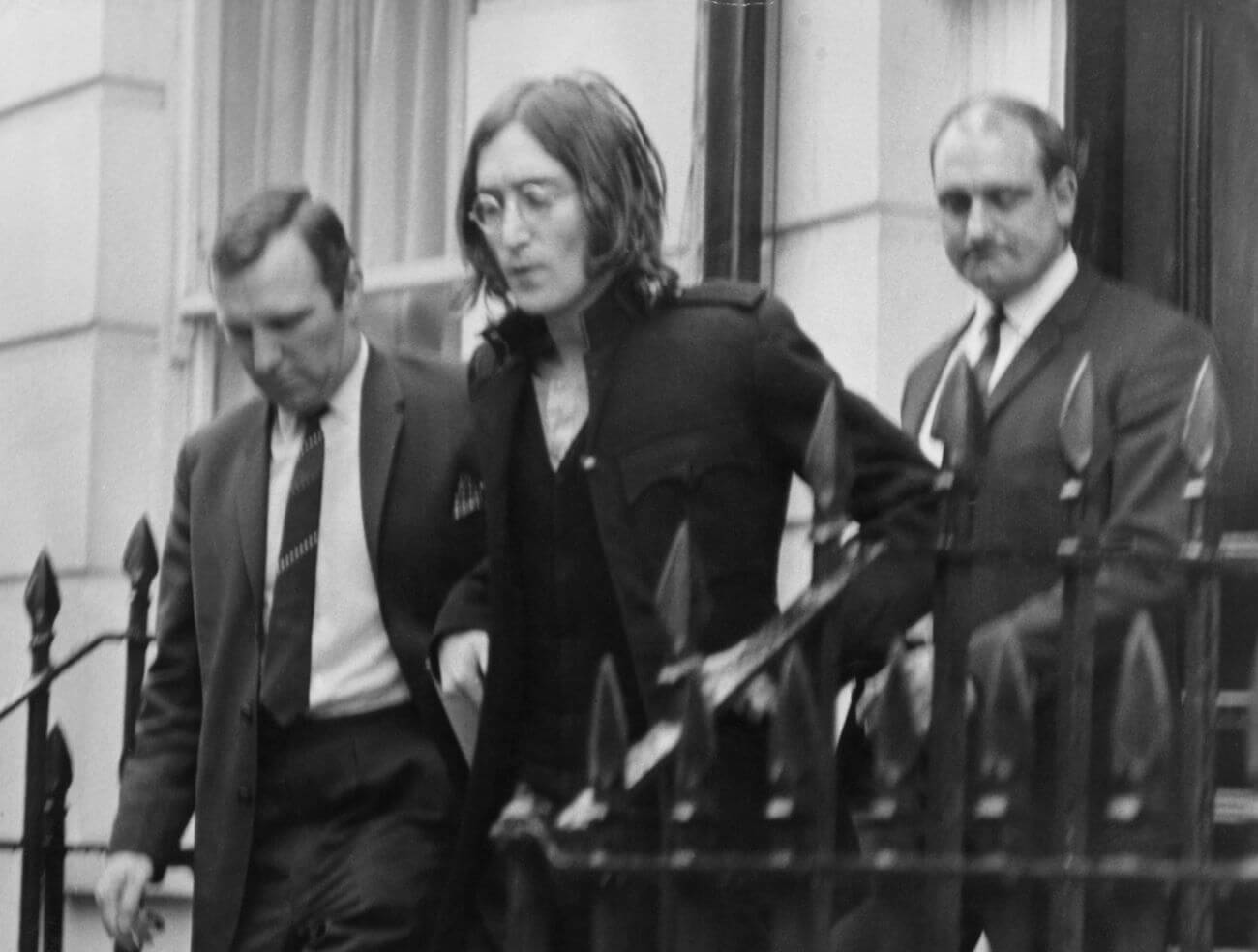 A black and white picture of two detectives leading John Lennon down steps outside his apartment. 