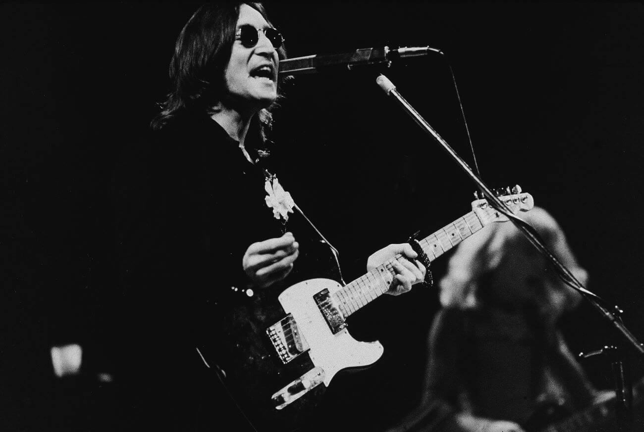 A black and white image of John Lennon wearing sunglasses, holding a guitar, and singing into a microphone.