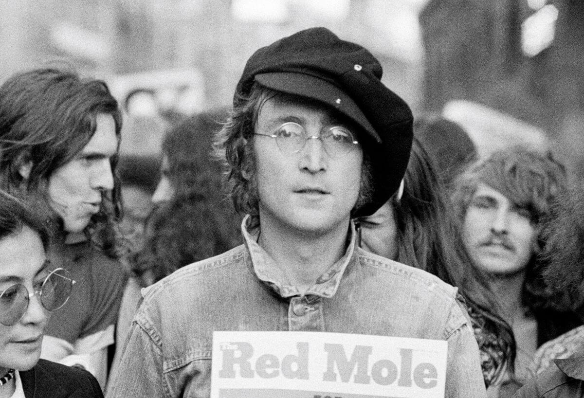 A black and white picture of John Lennon wearing a hat and holding a copy of The Red Mole in a crowd.