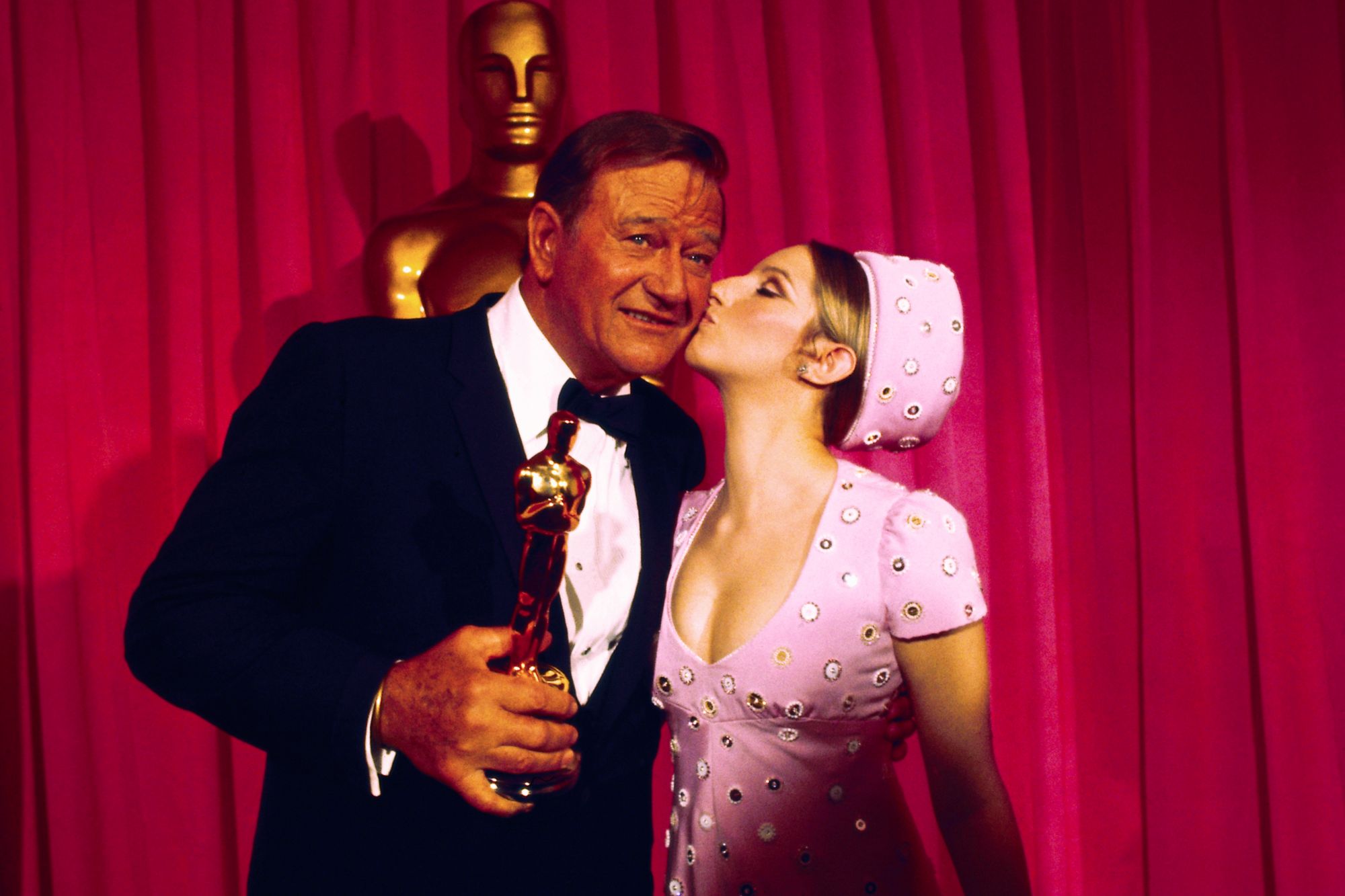 John Wayne and Barbra Streisand at the 1970 Oscars. She's kissing him on the cheek, while he holds his Academy Award. They're standing in front of an Oscar statue and red curtains.