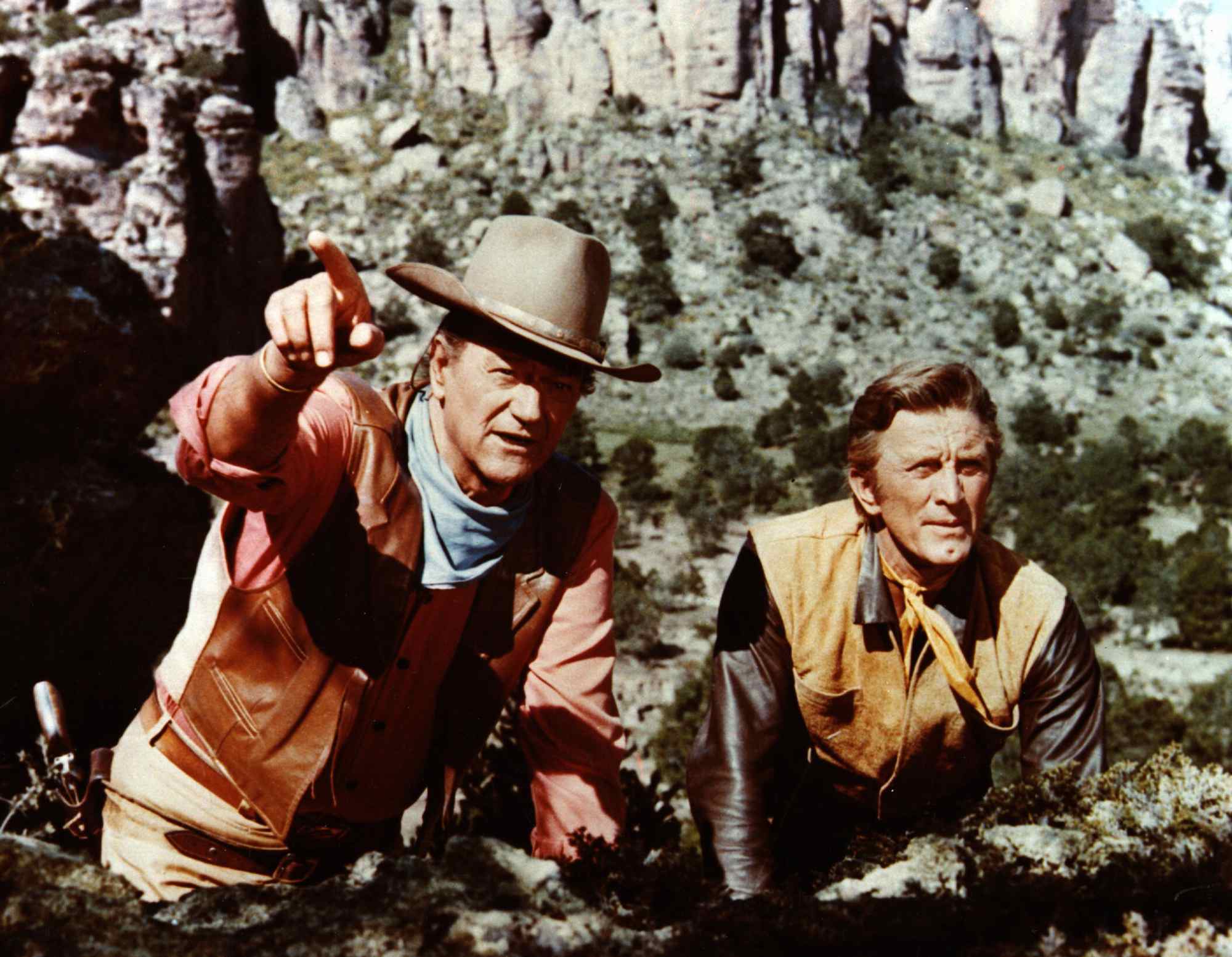 John Wayne and Kirk Douglas wearing Western costumes. Wayne is holding out his finger, pointing.