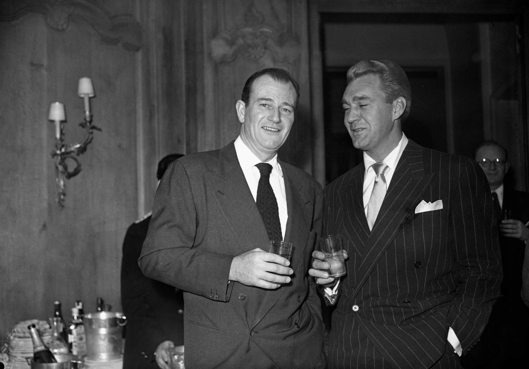 John Wayne having a drink with Forrest Tucker in a black-and-white picture. They're wearing suit and ties.