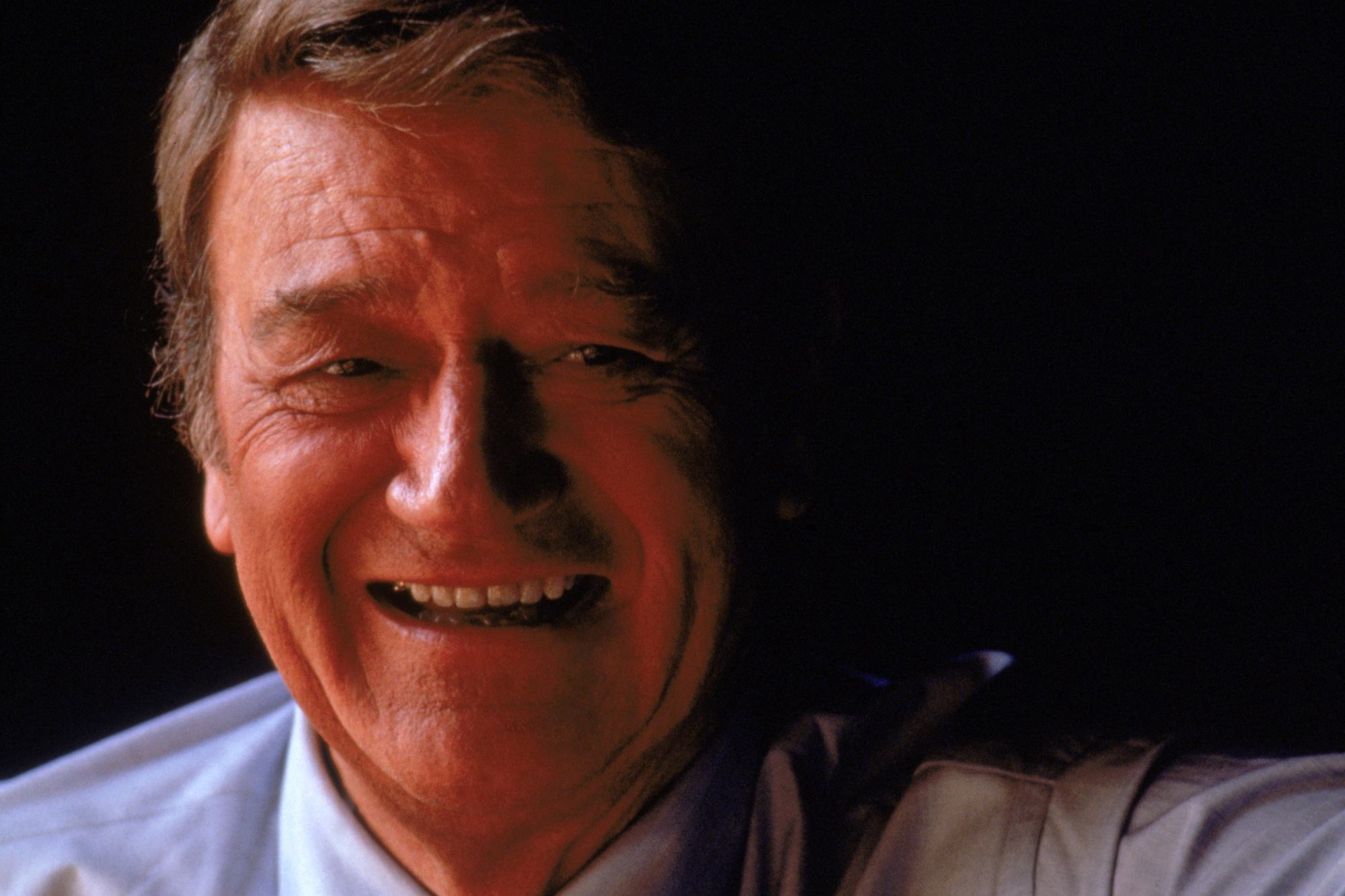 John Wayne, who married 3 women. A portrait picture with Wayne smiling and wearing a suit.