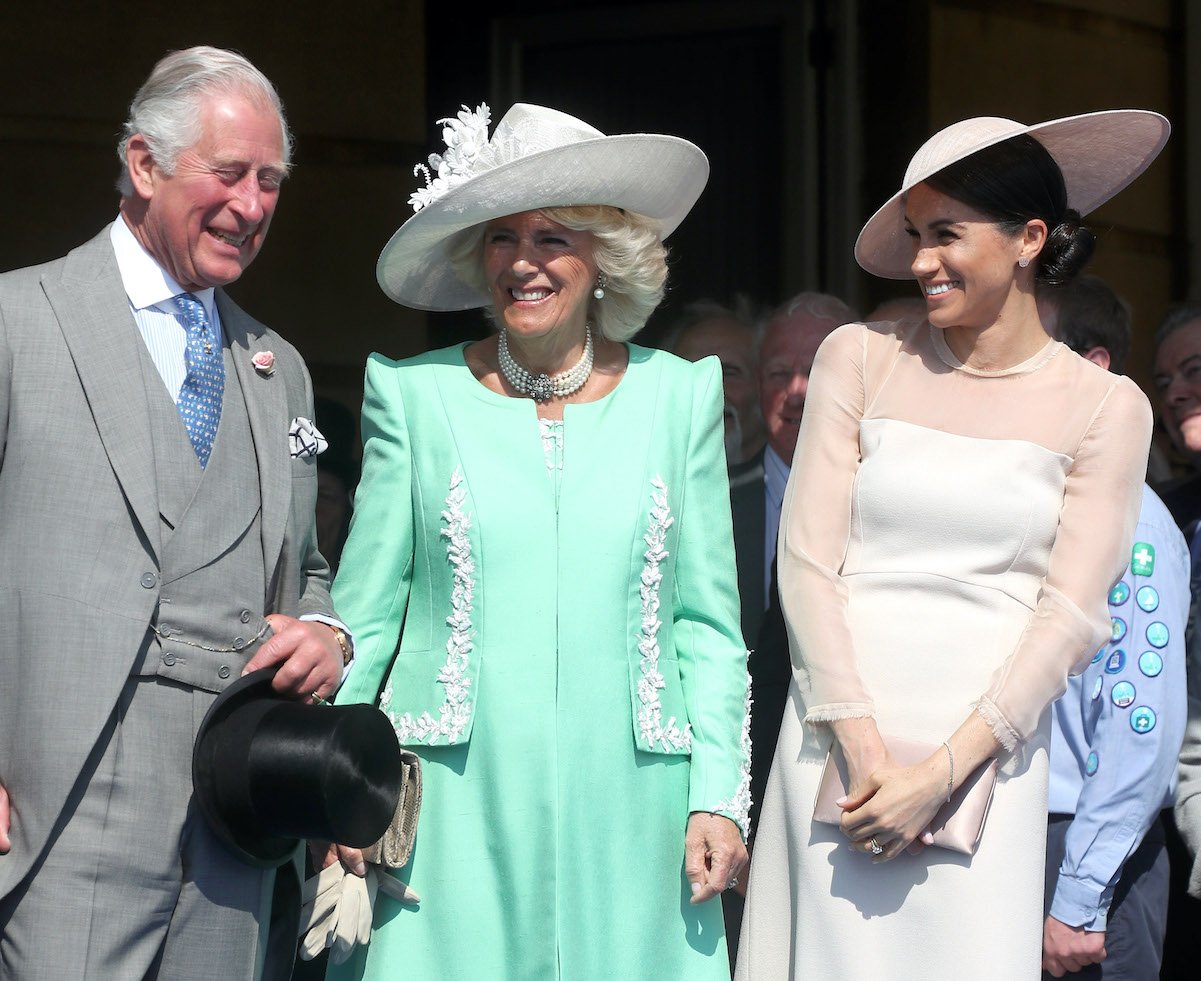 King Charles III stands with Camilla Parker Bowles and Meghan Markle. Charles reportedly is glad Meghan won't attend his coronation.