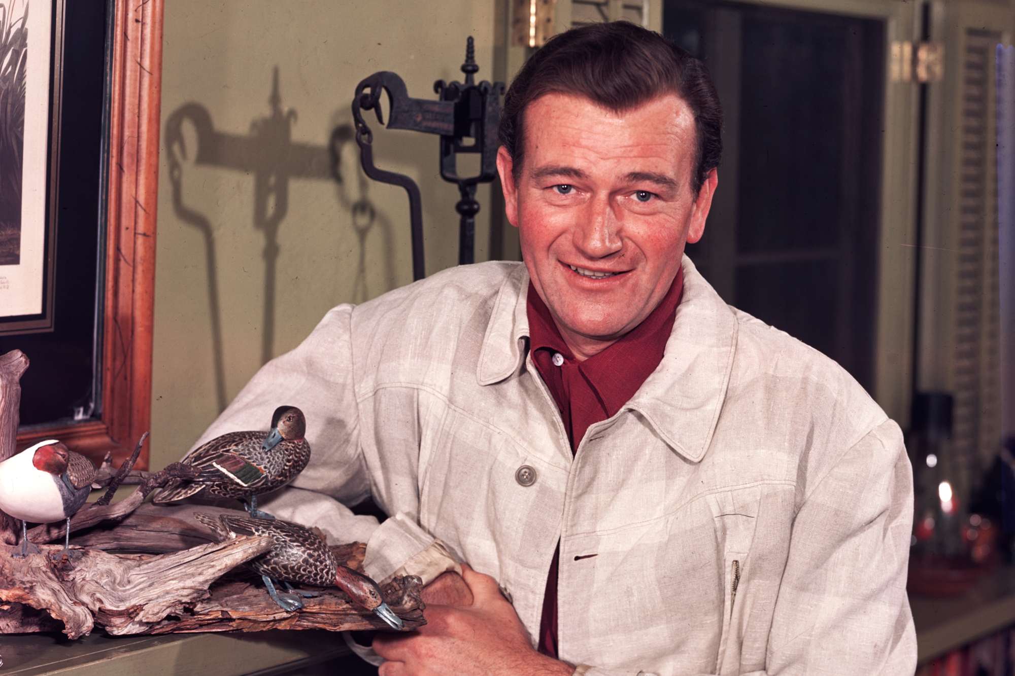 Life portrait of John Wayne leaning against a mantel next to a wooden duck statue.