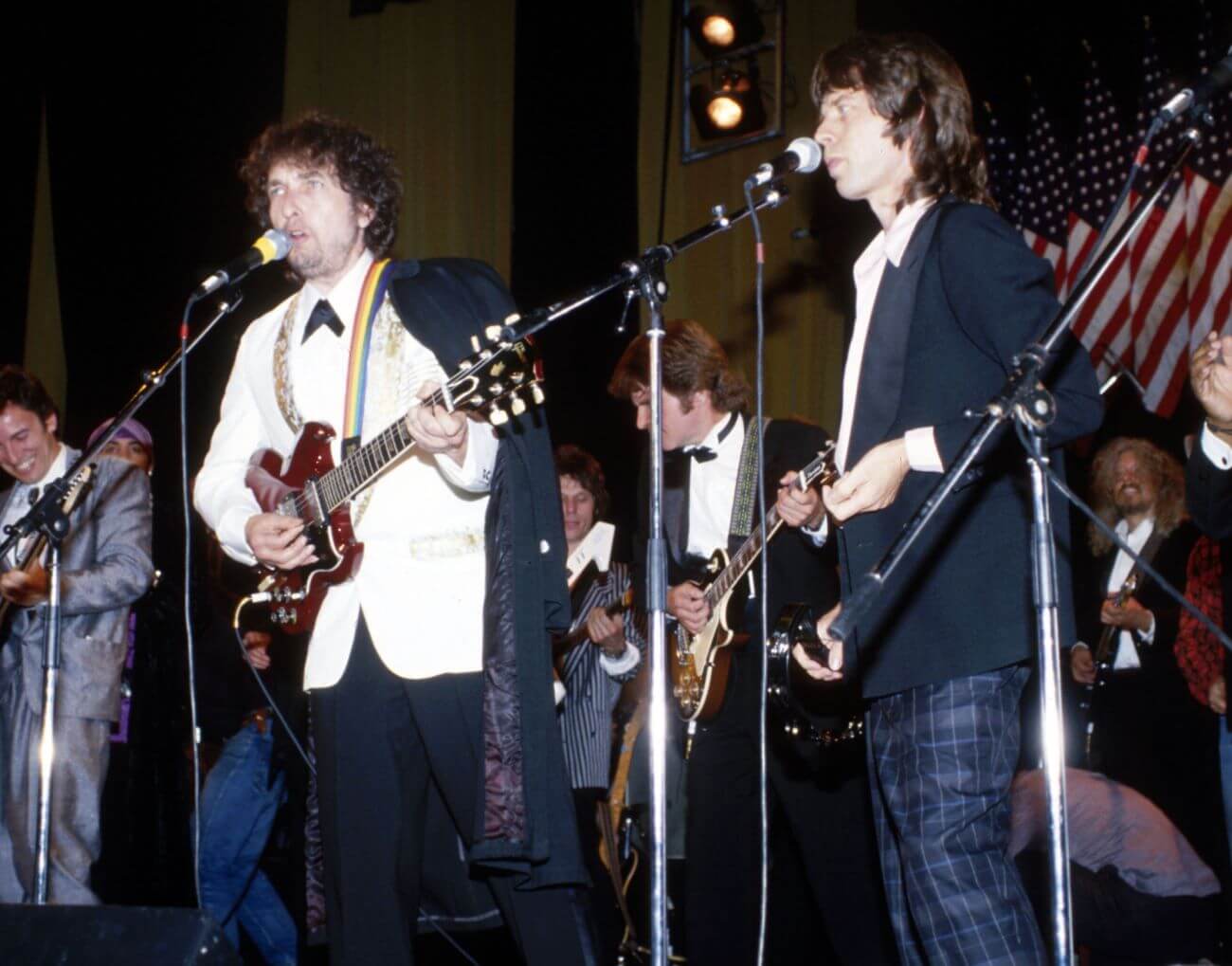 Bob Dylan plays guitar and sings into a microphone. Mick Jagger stands in front of a microphone.