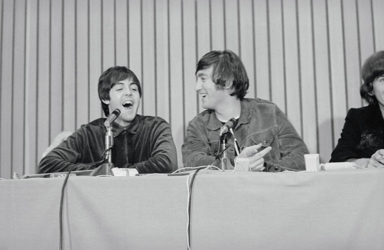 A black and white picture of Paul McCartney and John Lennon laughing while sitting in front of microphones at a table.
