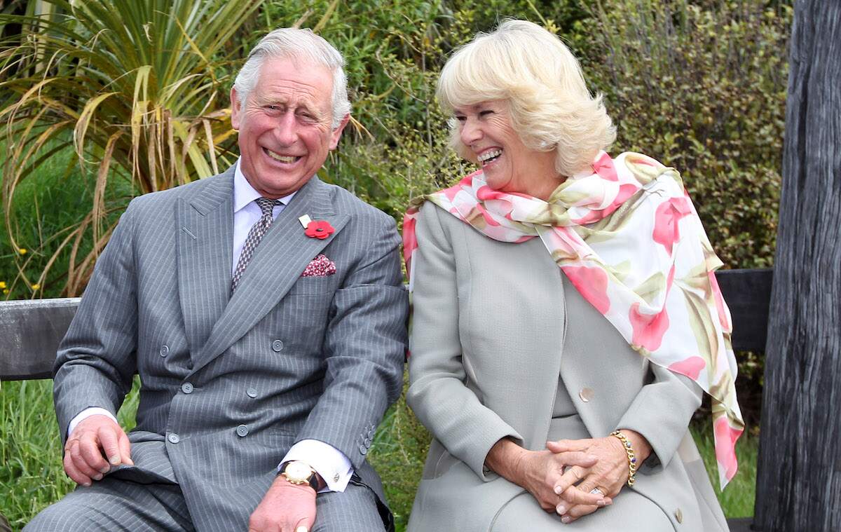 Married couple Prince Charles, Prince of Wales and Camilla, Duchess of Cornwall laugh together while sitting on a bench
