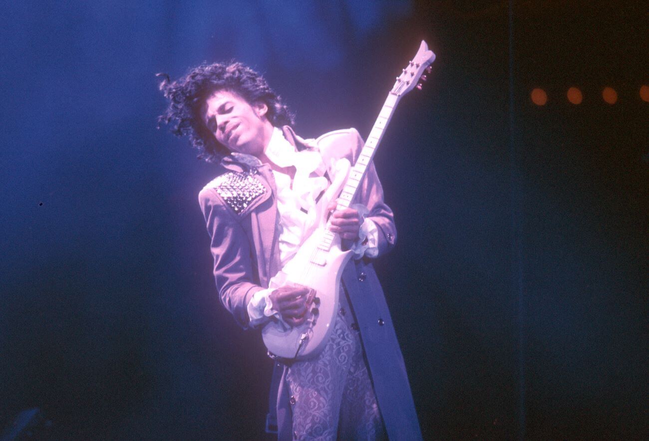 Prince wears a purple jacket and plays guitar.