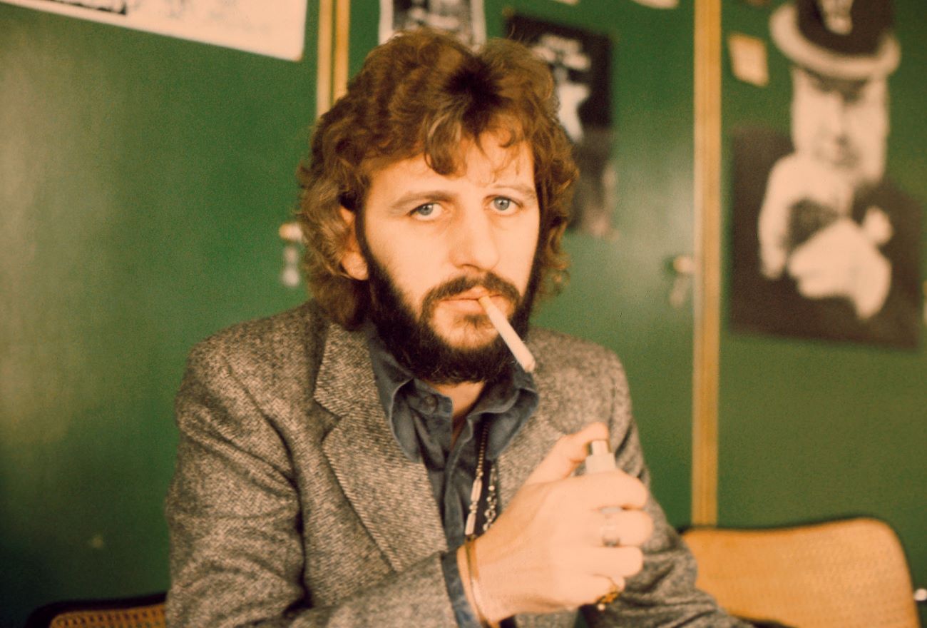 Ringo Starr sits in front of green walls a lights a cigarette.
