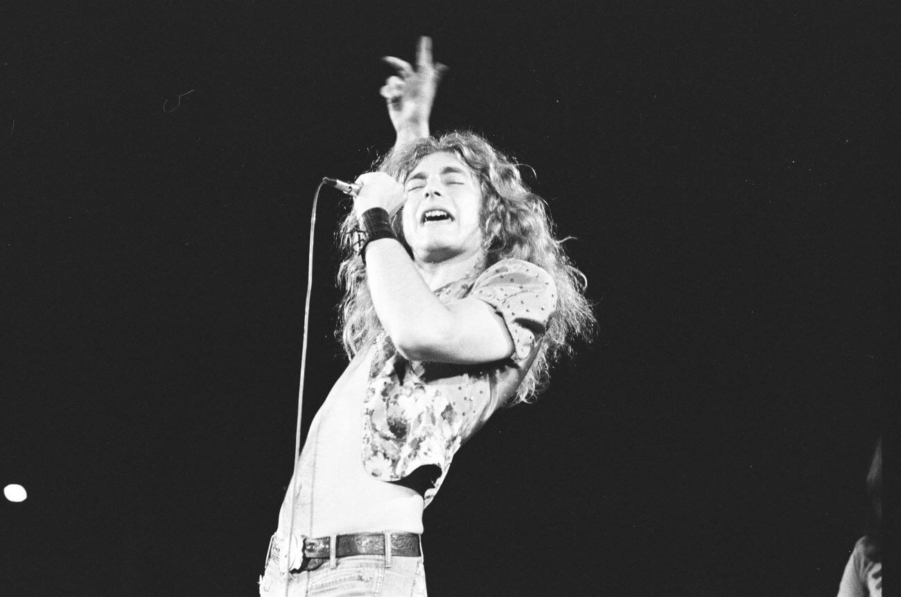A black and white picture of Robert Plant leaning backward and holding his arm up while singing into a microphone.