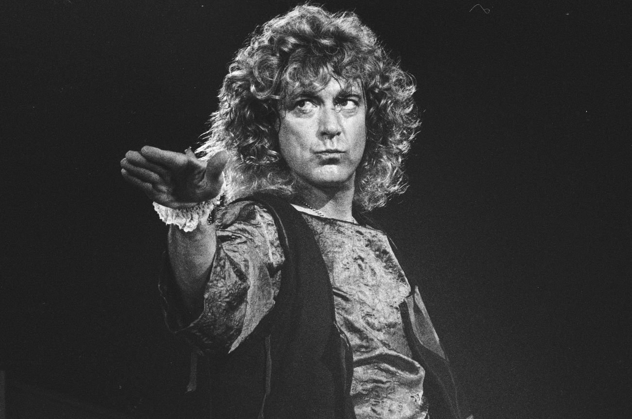 A black and white picture of Robert Plant wearing a shirt and a vest and holding one arm out.