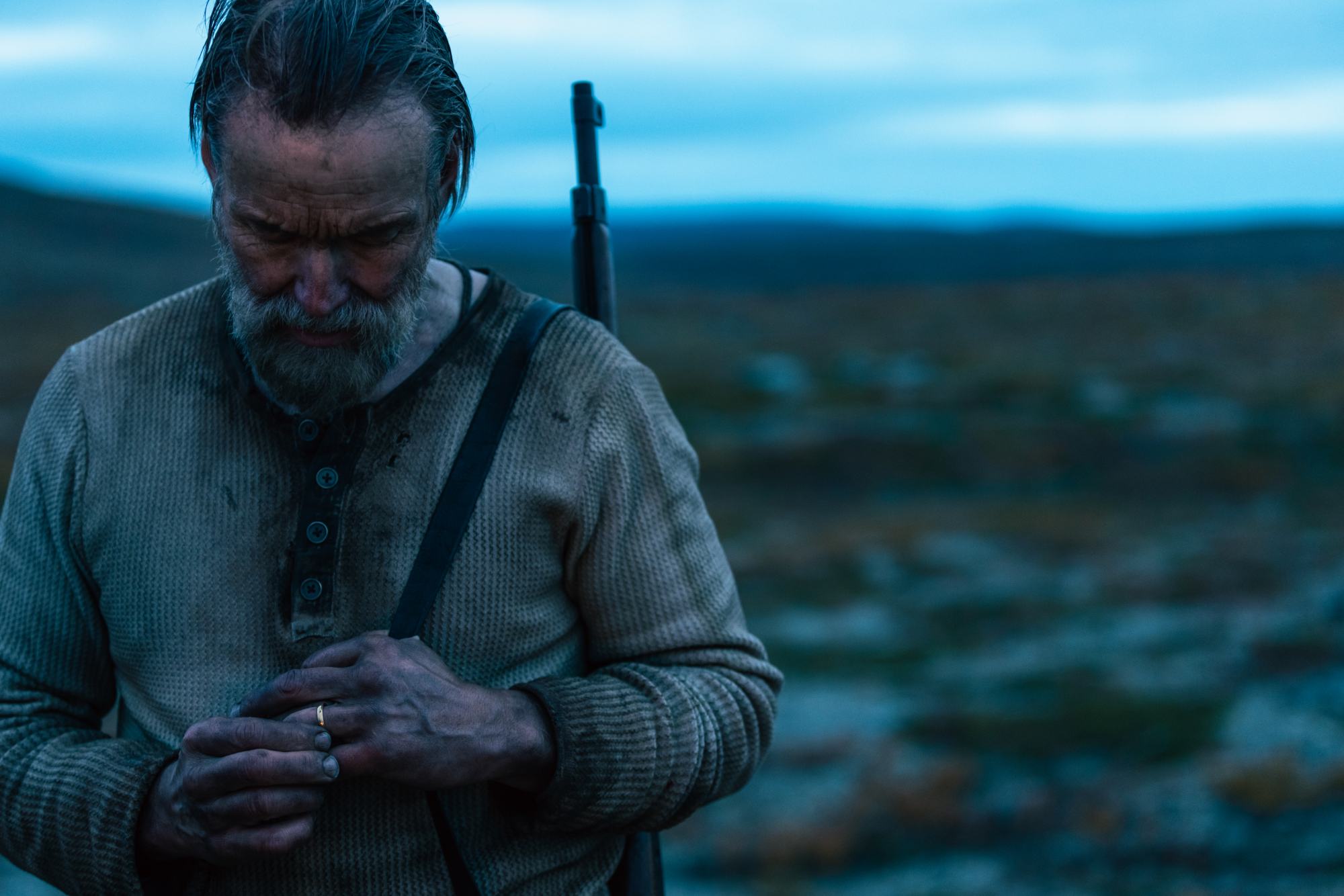 'Sisu' Jorma Tommila as Aatami Korpi wearing dirty clothes with a gun strapped on his back. He's playing with the gold wedding band on his finger.