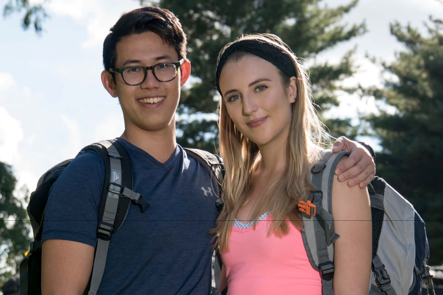 Henry Zhang and Evan Lynyak pose for promotional photos for 'The Amazing Race 30' on CBS. Henry wears a dark blue shirt and glasses. Evan wears a light pink tank top and black headband.