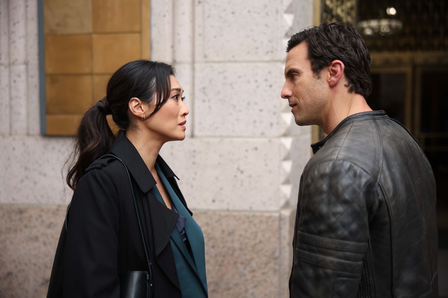 Catherine Haena Kim and Milo Ventimiglia, in character as Emma and Charlie in 'The Company You Keep,' share a scene in the show. Emma wears a black coat over a teal suit. Charlie wears a black leather jacket.