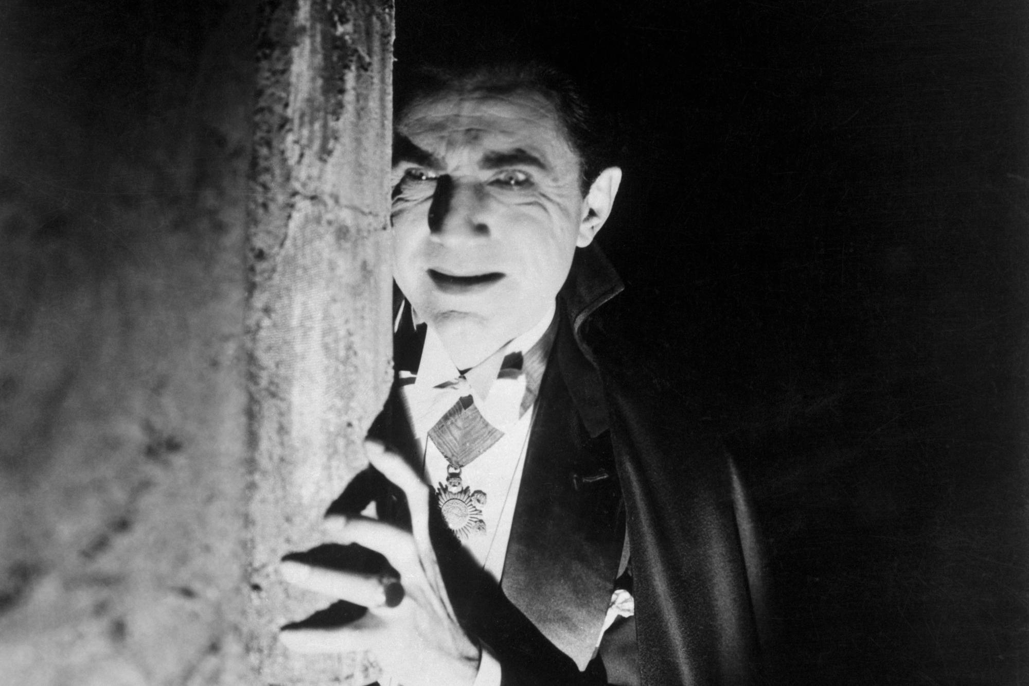 Vampire movies 'Dracula' Bela Lugosi as Dracula hiding behind a wall, with his hand resting on it. He has a devilish smile.