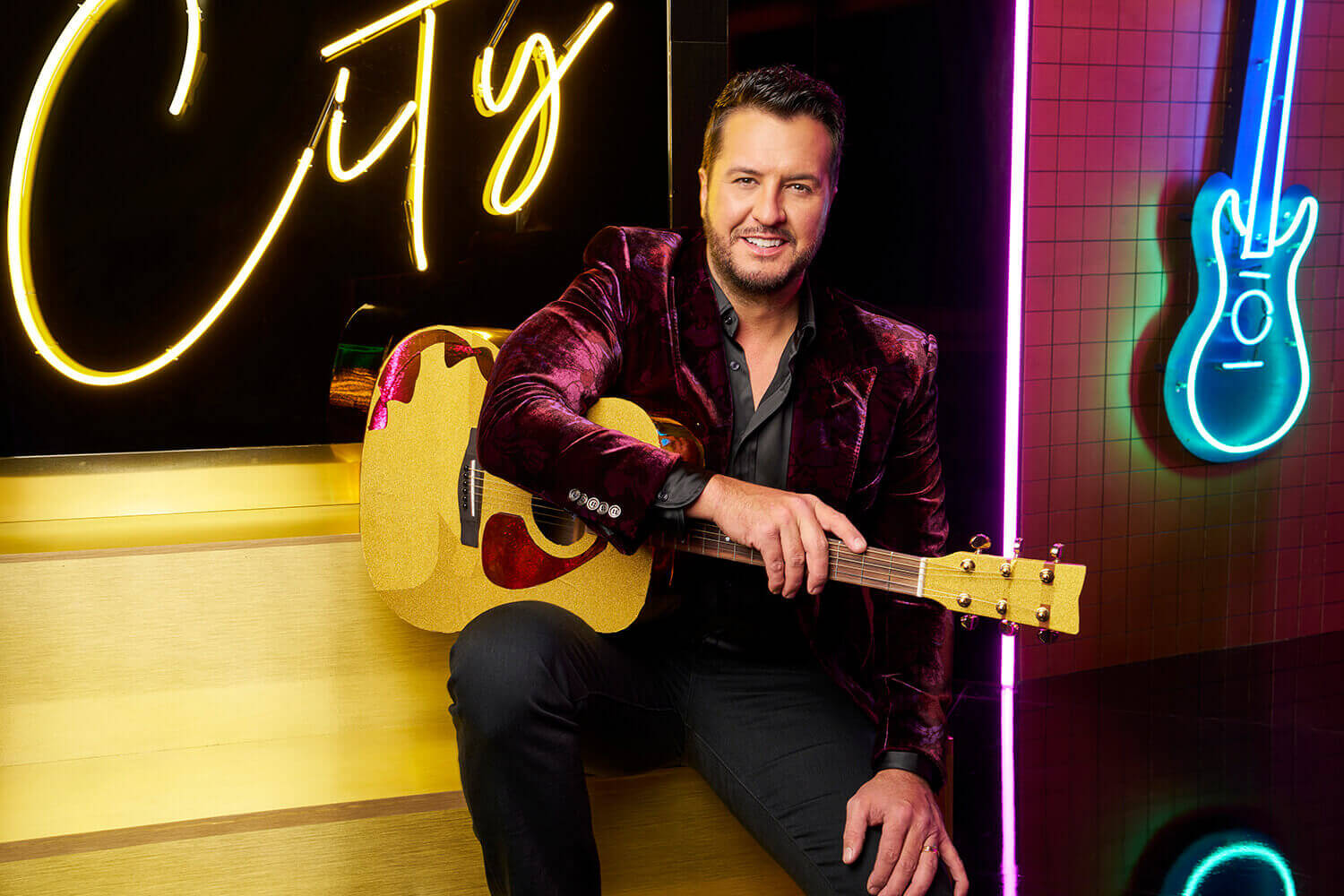 American Idol judge Luke Bryan sits on a yellow bench with a guitar on his knee and smiles