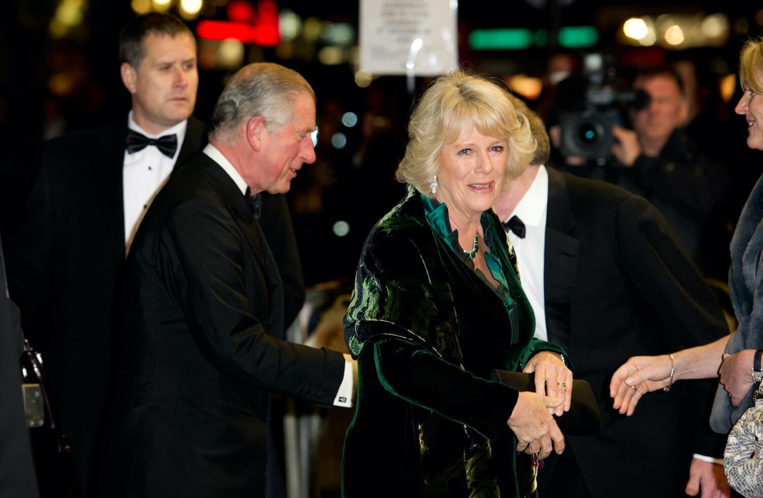 King Charles dressed in a tux walks behind Camilla Parker Bowles wearing a green gown