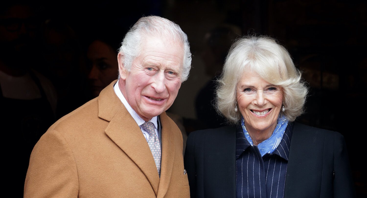 King Charles wears a brown coat and stands next to Camilla Parker Bowles dressed in a striped outfit and coat, smiling