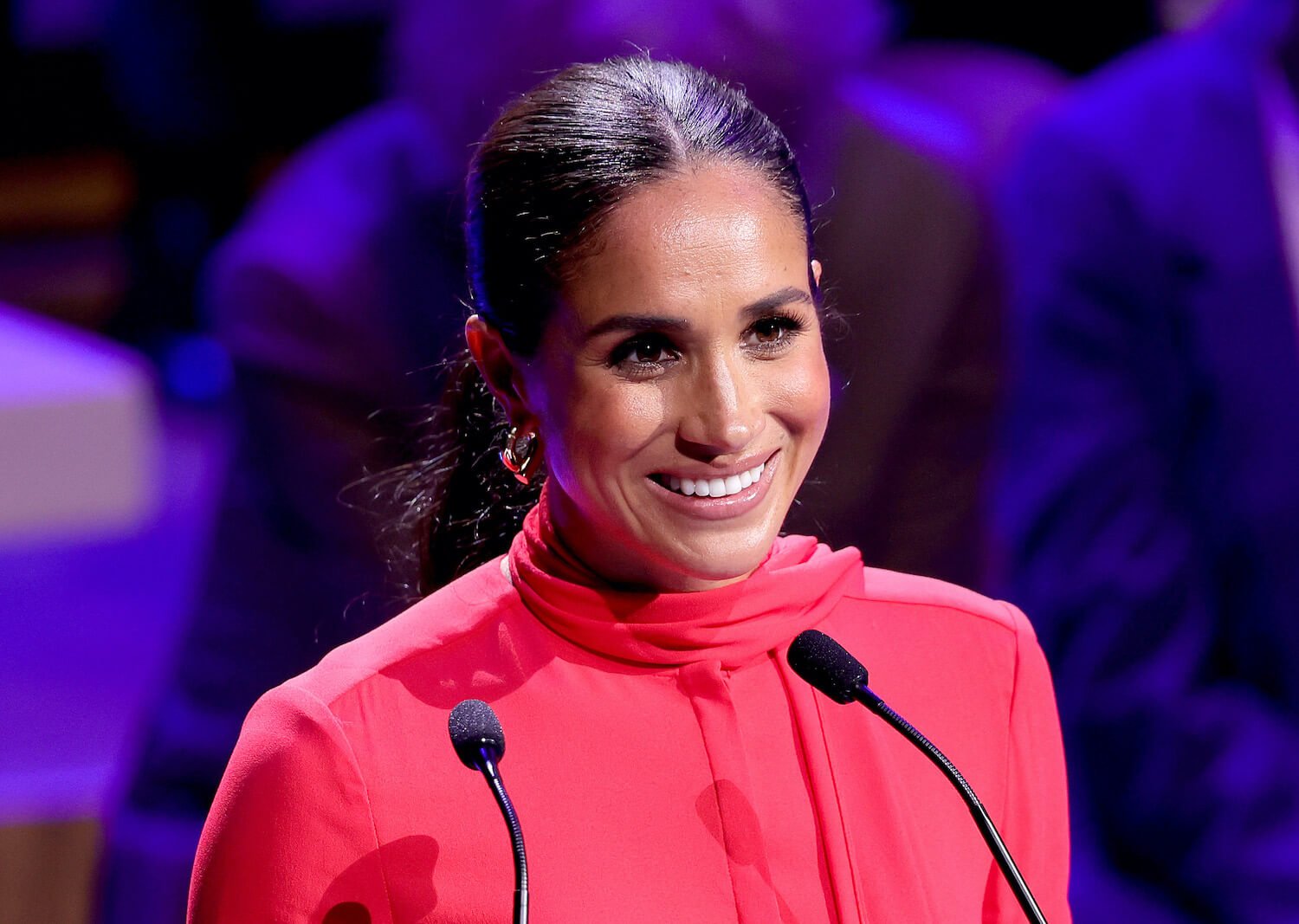 Meghan Markle wears a red top and smiles with her hair pulled back
