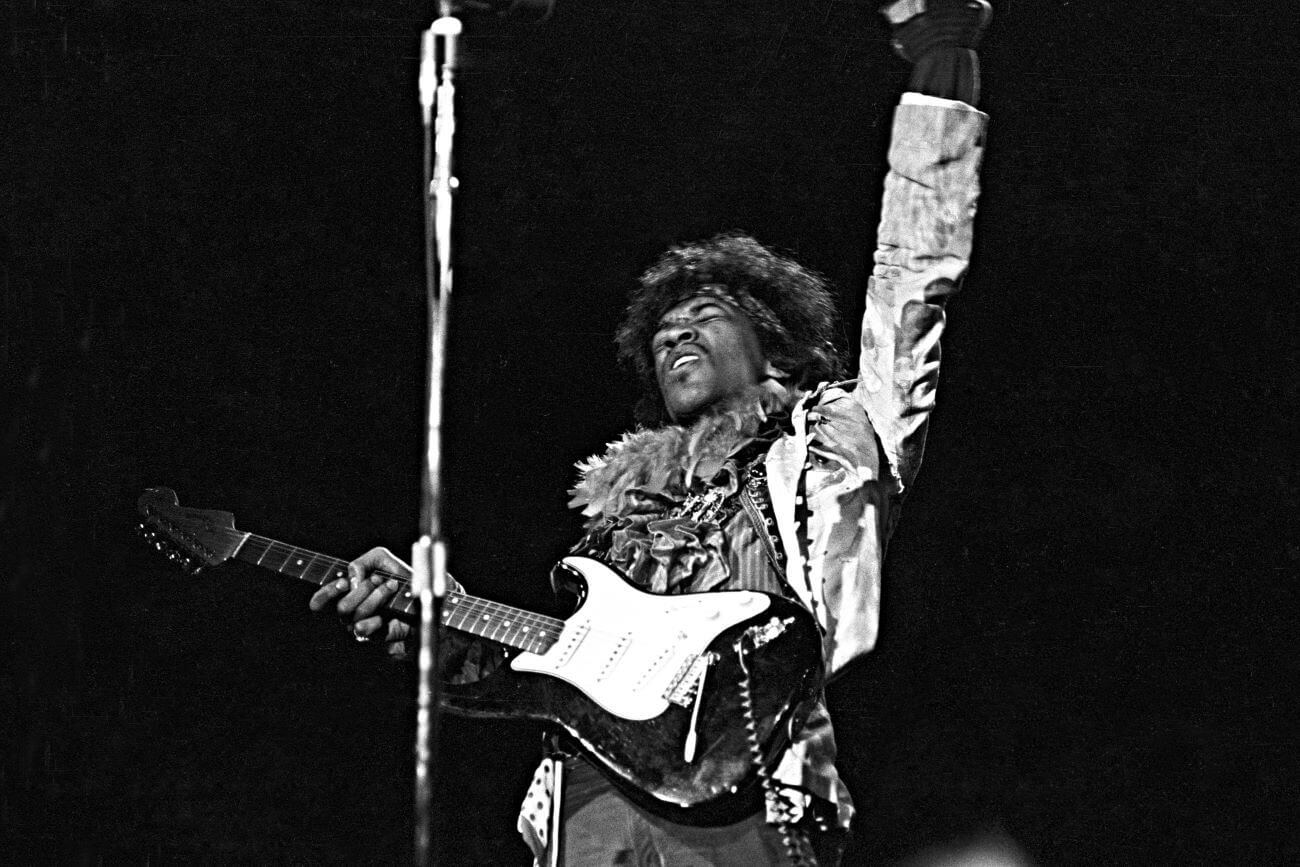 A black and white picture of Jimi Hendrix holding a guitar and lifting his arm into the air during a music festival.