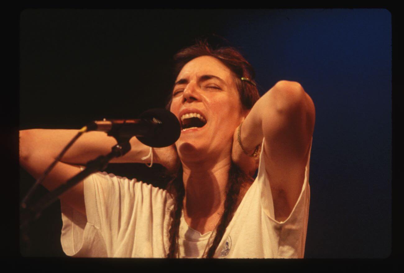 The musician Patti Smith holds her hands behind her head while she sings into a microphone.