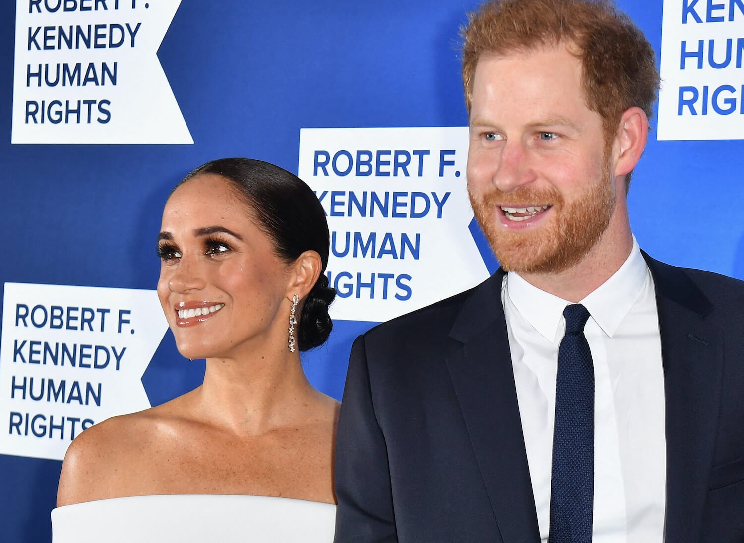 Prince Harry wears a dark suit and smile and smiles beside Meghan Markle wearing a white off the shoulder top and smiling