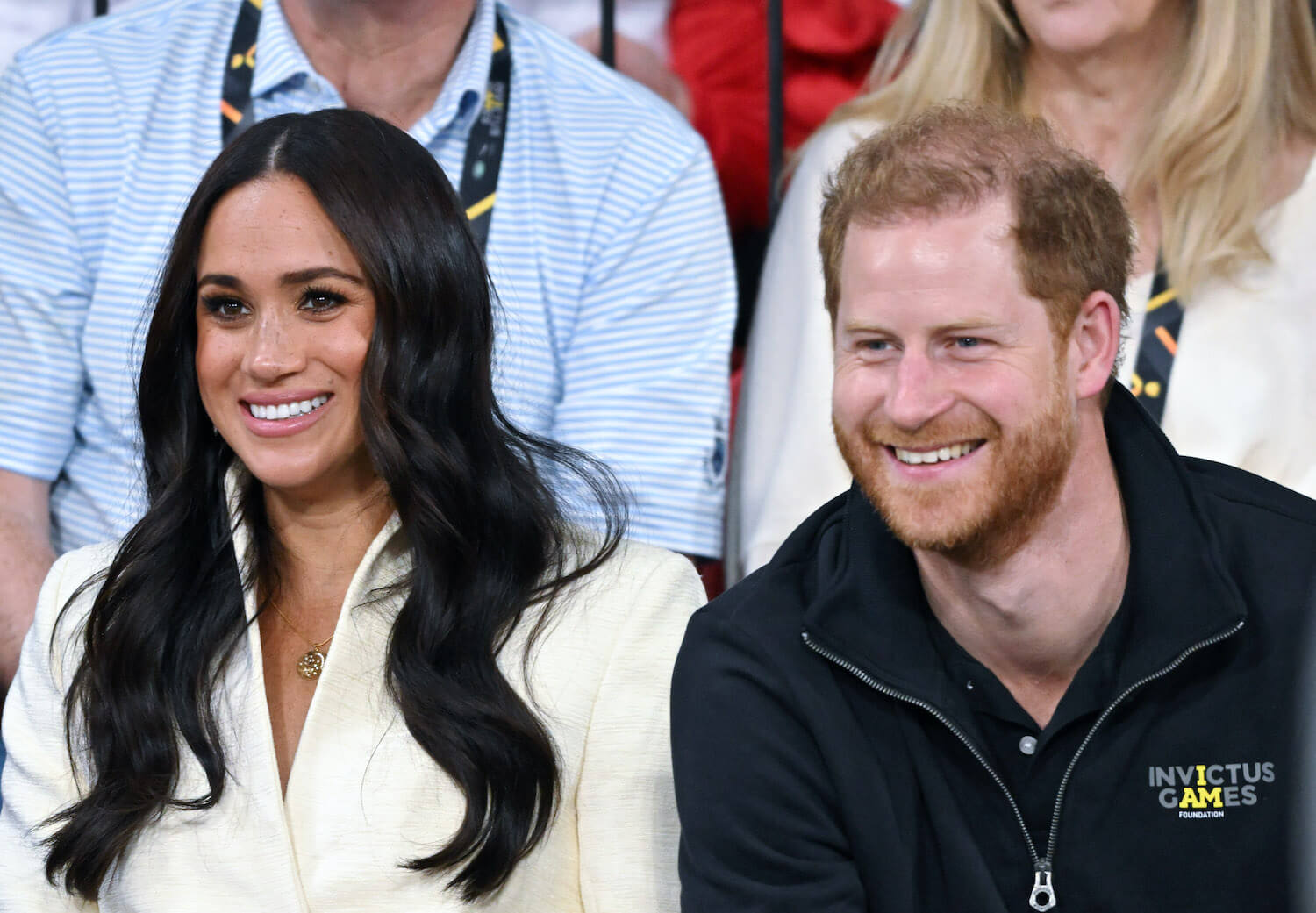 Meghan Markle wears a white top and smiles next to Prince Harry smiling while wearing a dark zip up shirt
