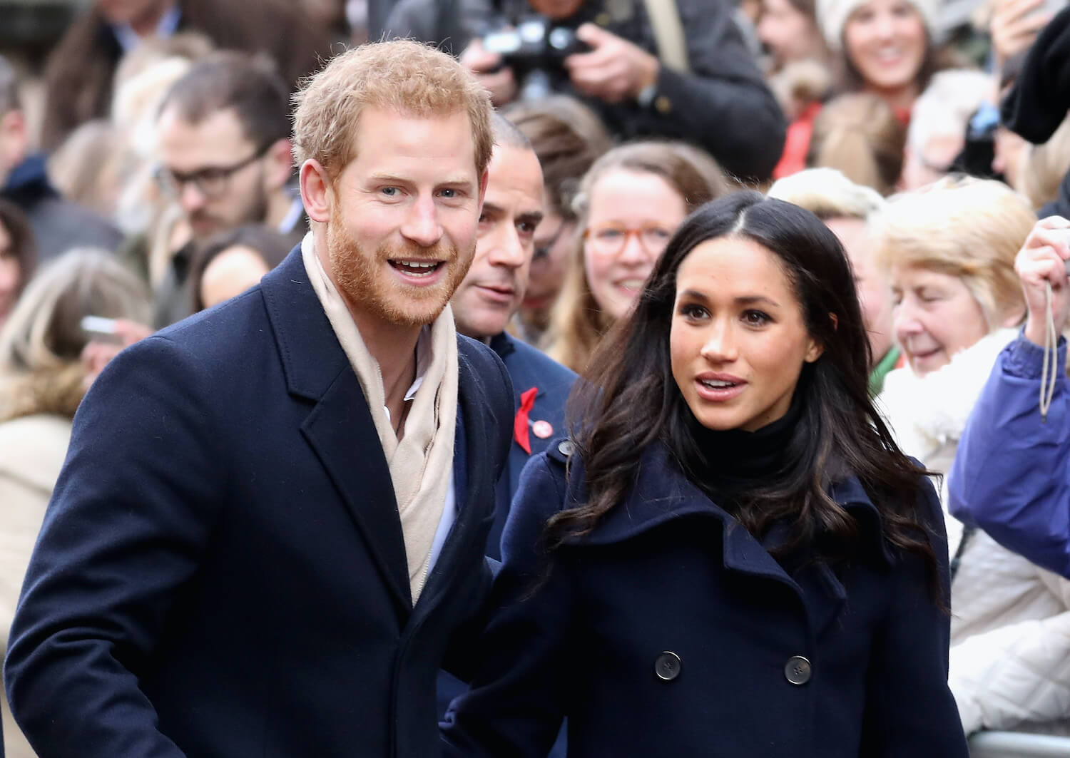 Prince Harry grins wearing a coat alongside Meghan Markle who looks off while wearing a dark coat