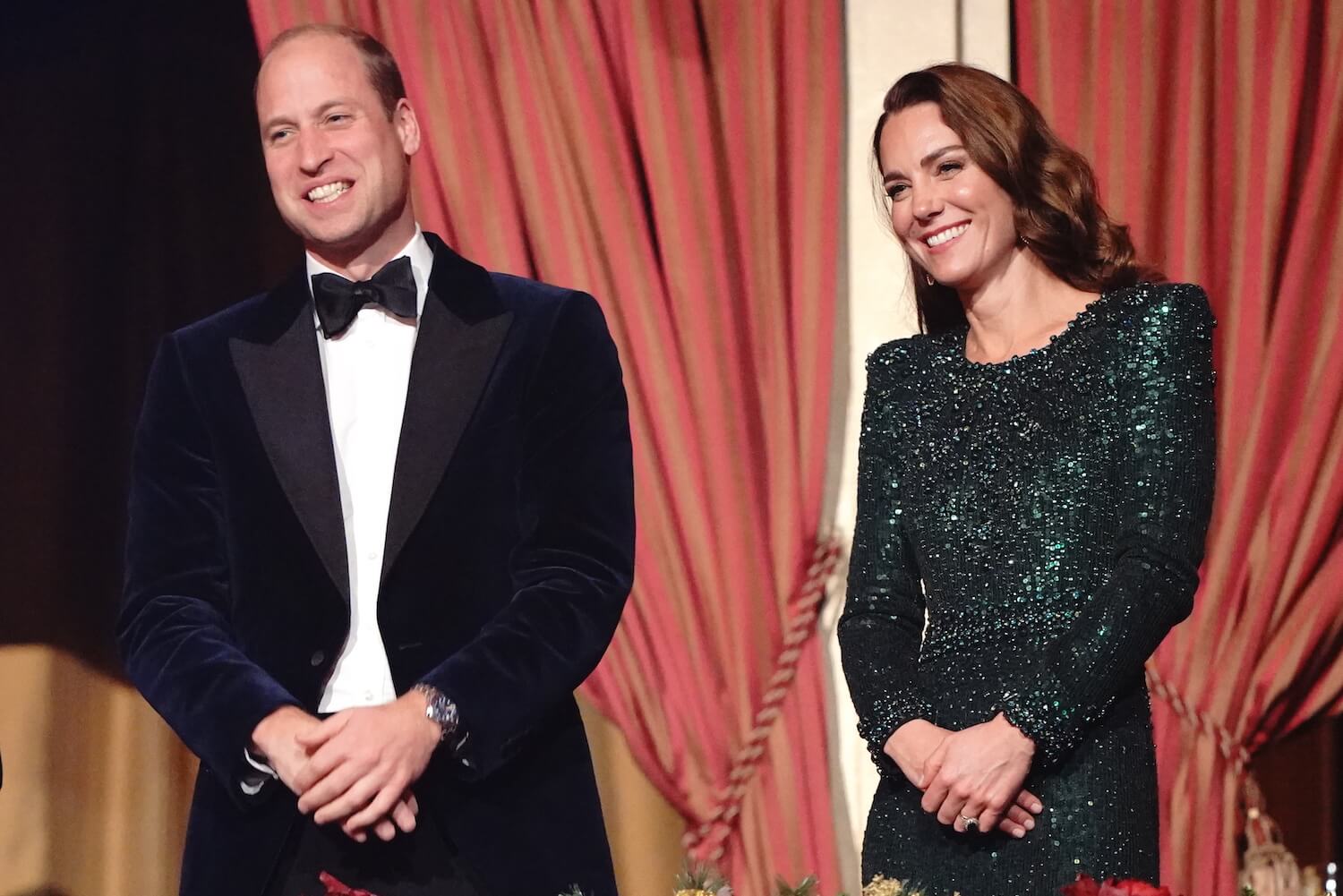 Prince William wears a black tux and smiles with Kate Middleton standing by him, wearing a green gown and smiling