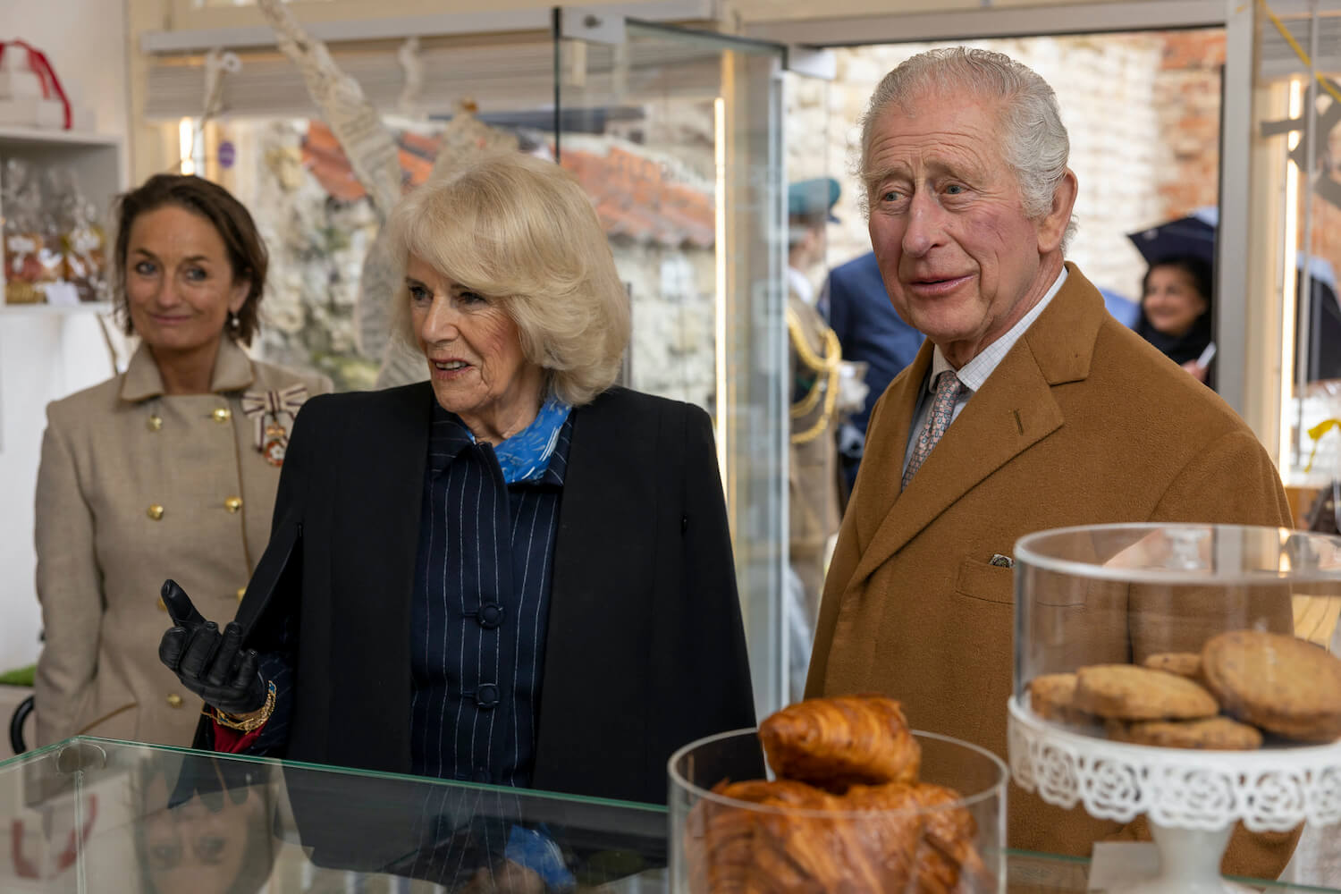King Charles wearing a brown coat and Queen Camilla wearing a dark coat stand at a bakery counter