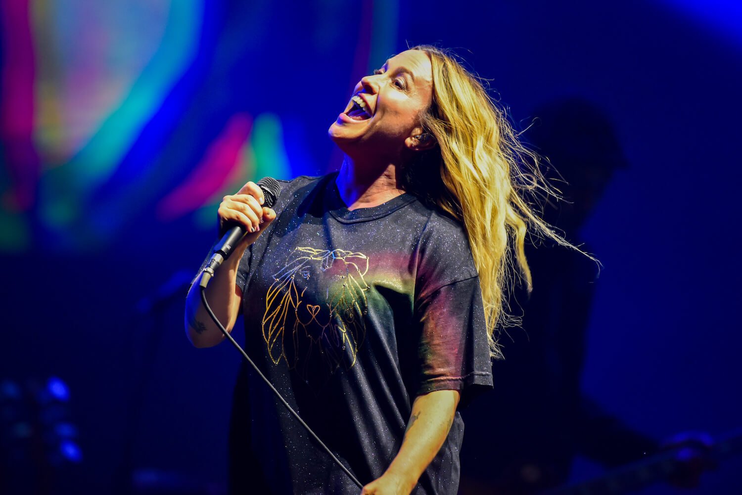 Alanis Morissette singing into a microphone on stage in a T-shirt against a blue background. Alanis Morissette is a guest judge on 'American Idol' 2023.