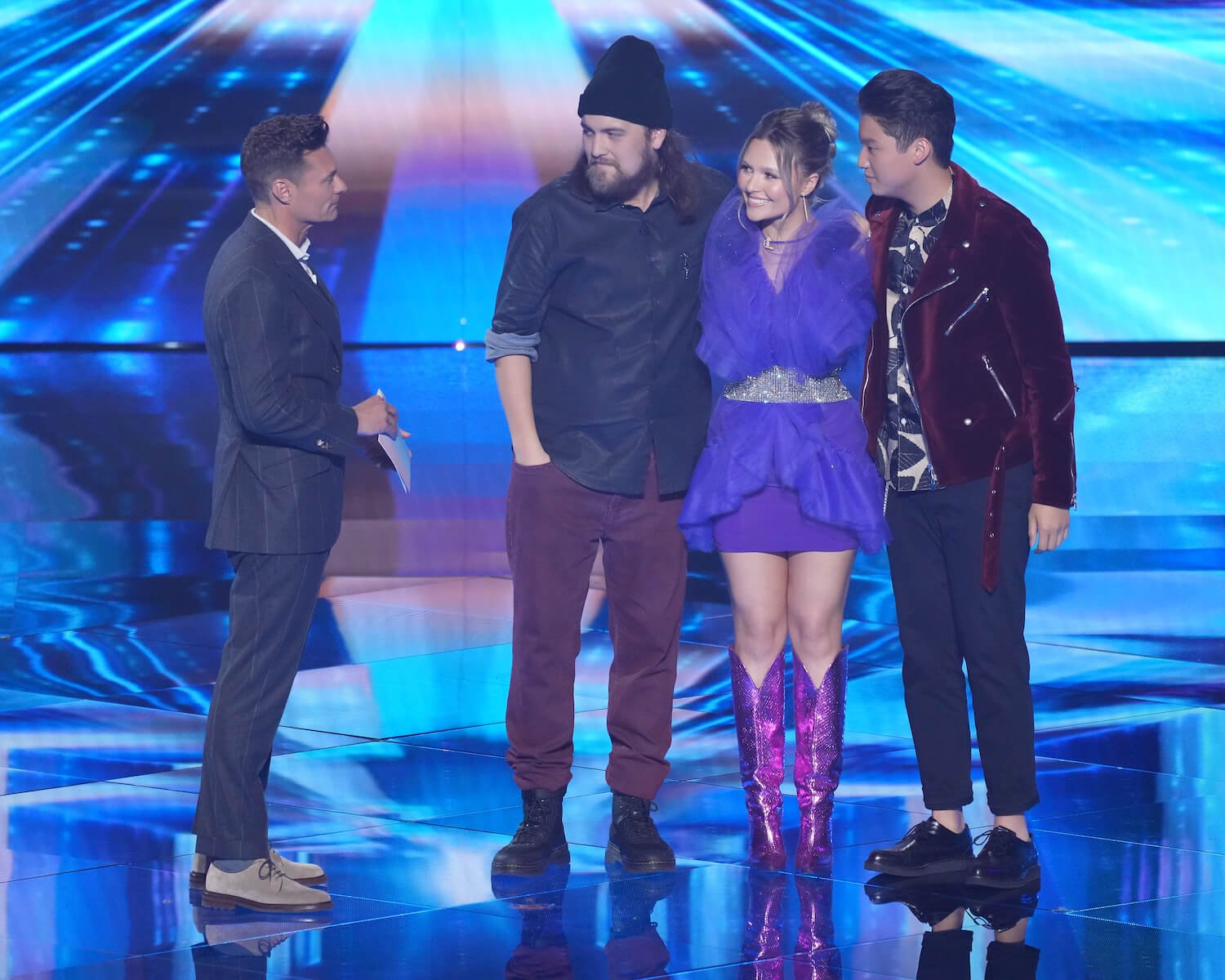 Ryan Seacrest talking to three contestants huddled together on stage in 'American Idol' 2023