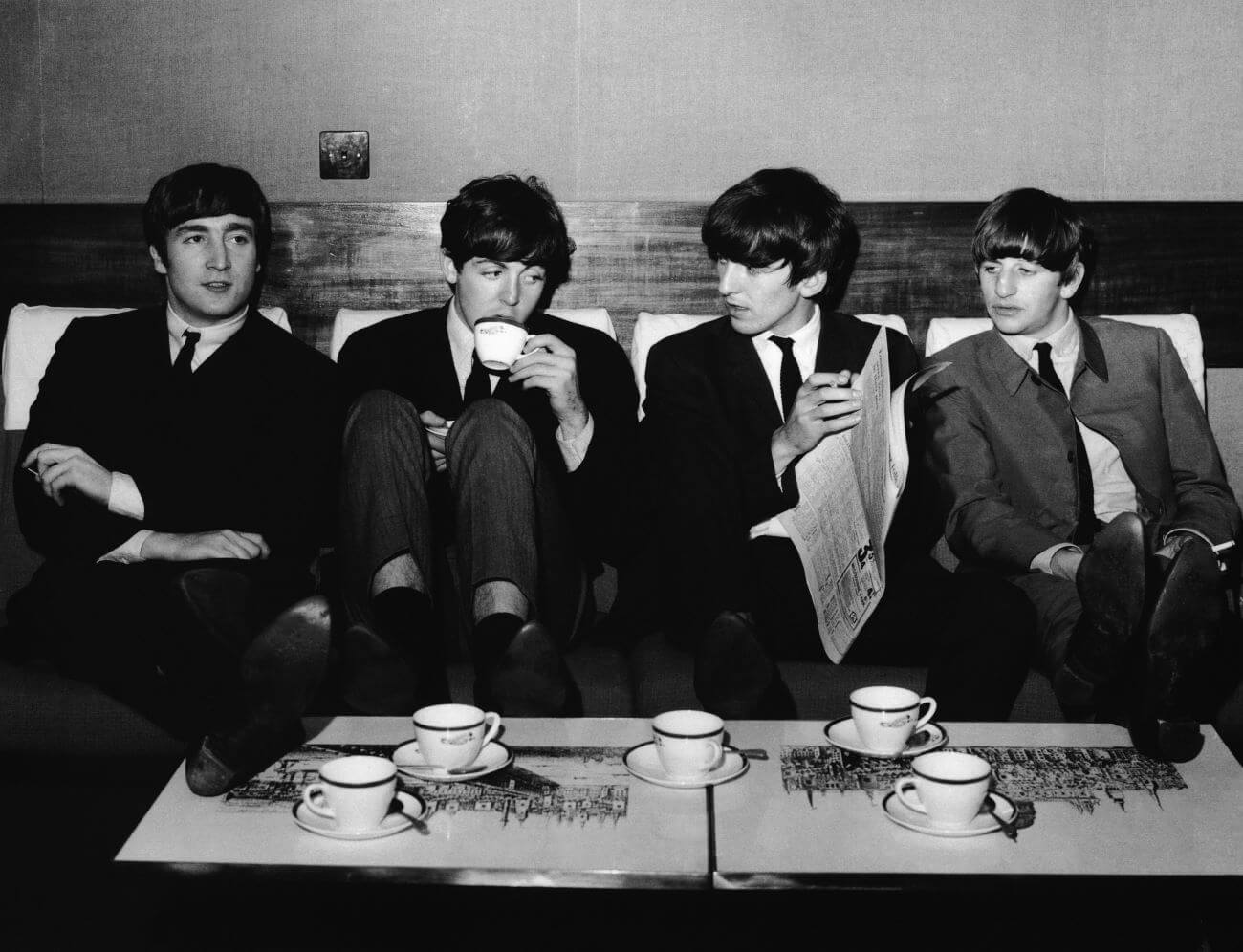 A black and white picture of John Lennon, Paul McCartney, George Harrison and Ringo Starr of The Beatles sitting on a couch in front of a table full of tea cups. Harrison holds a newspaper.