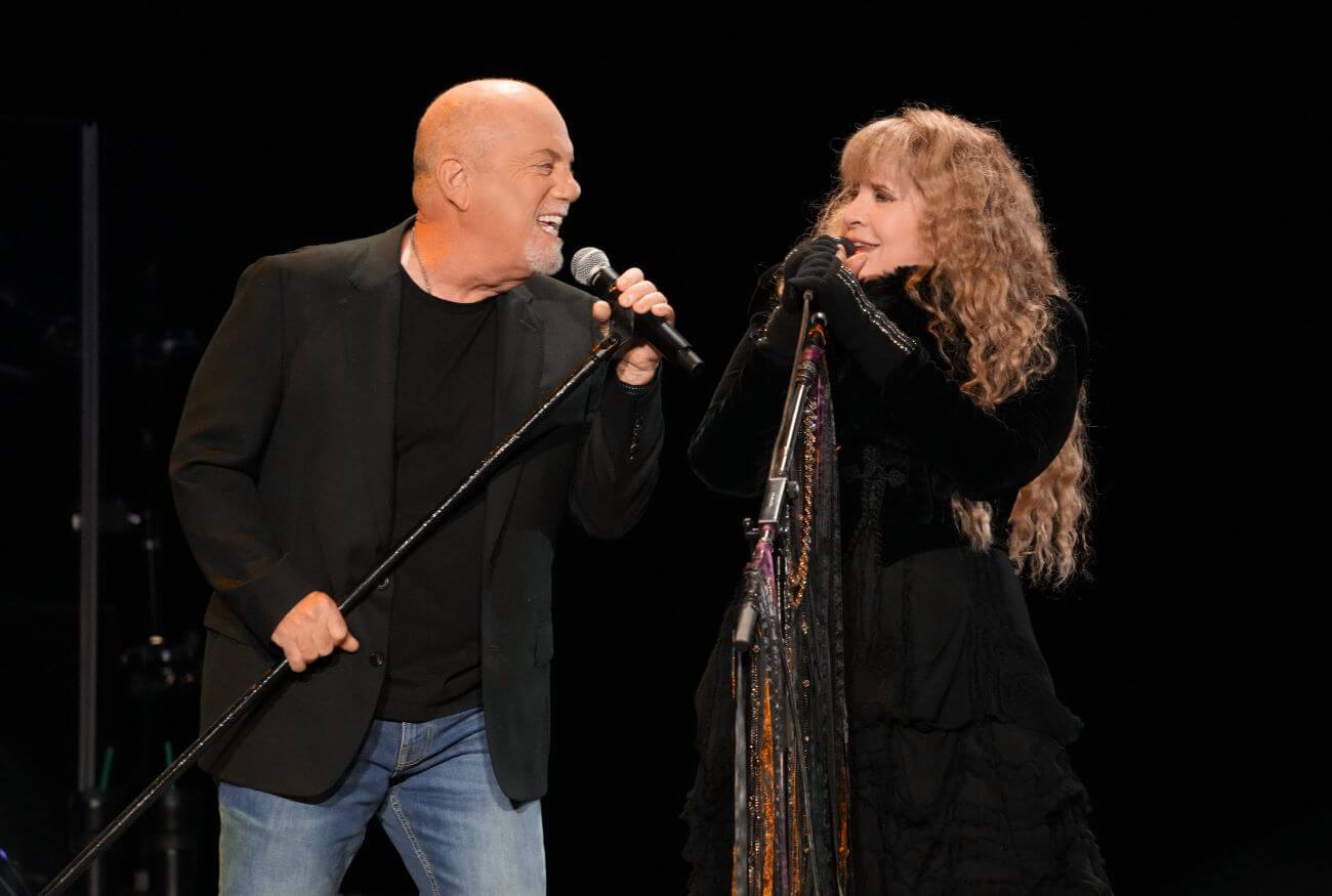 Billy Joel and Stevie Nicks both wear black and sing into microphones. 