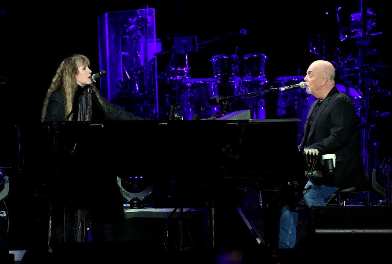 Stevie Nicks and Billy Joel sing into microphones while Joel plays the piano.