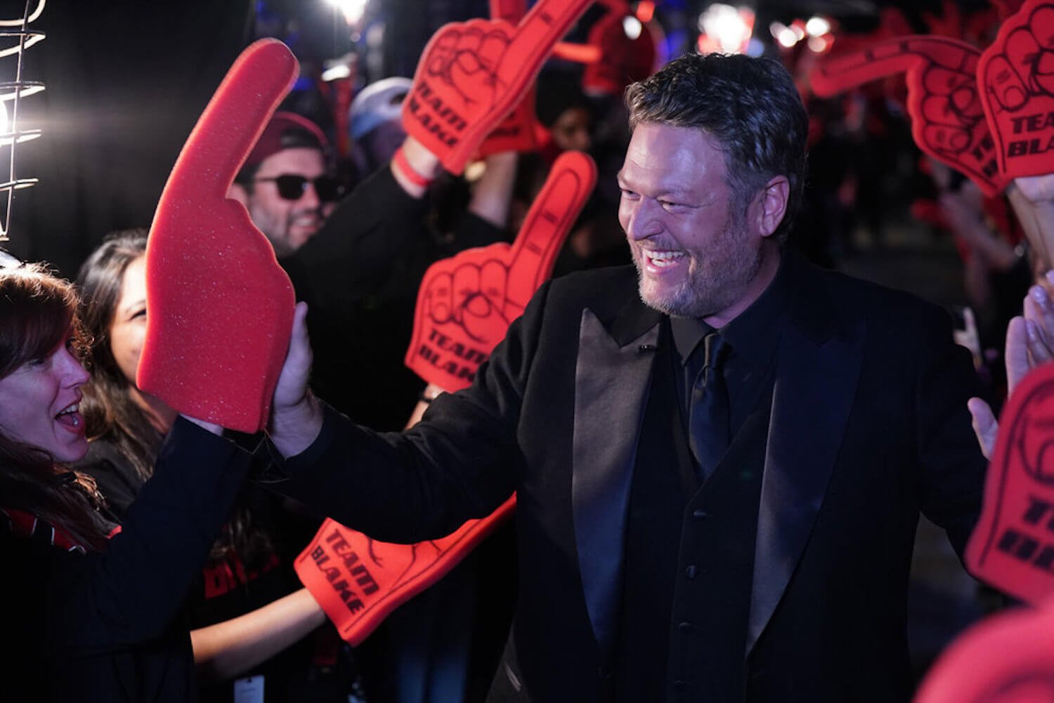 Blake Shelton from 'The Voice' laughing while surrounded by fans with foam fingers
