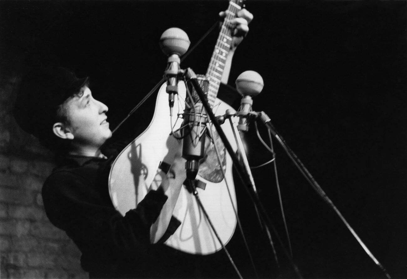 A black and white image of Bob Dylan wearing a hat and playing guitar in front of three microphones in a club in New York.