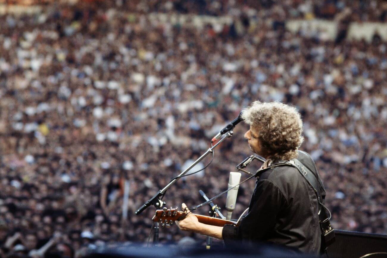 Bob Dylan plays guitar and stands in front of a microphone in front of a large group of fans. 