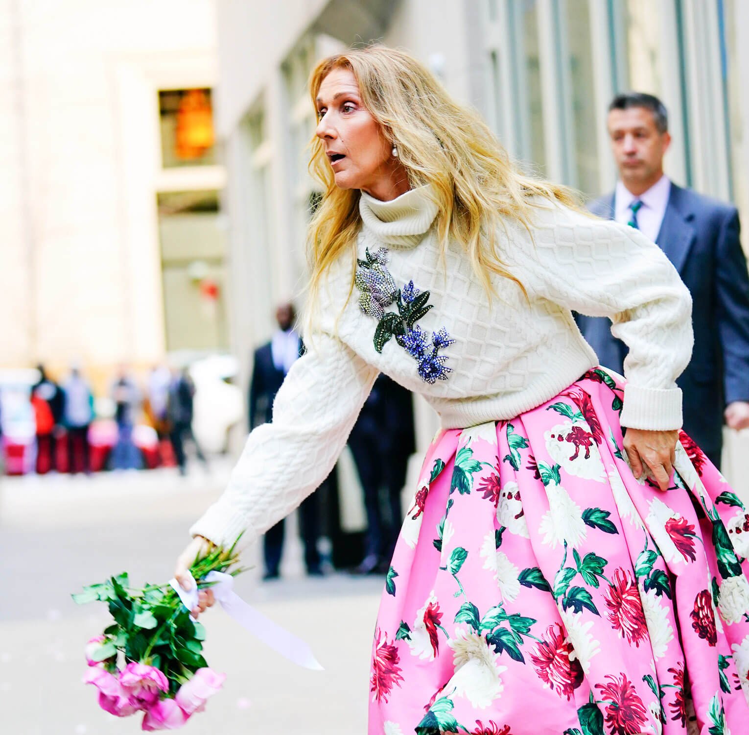 Celine Dion standing with her hand out holding roses on the street. She's wearing a white turtleneck sweater and a pink floral skirt.